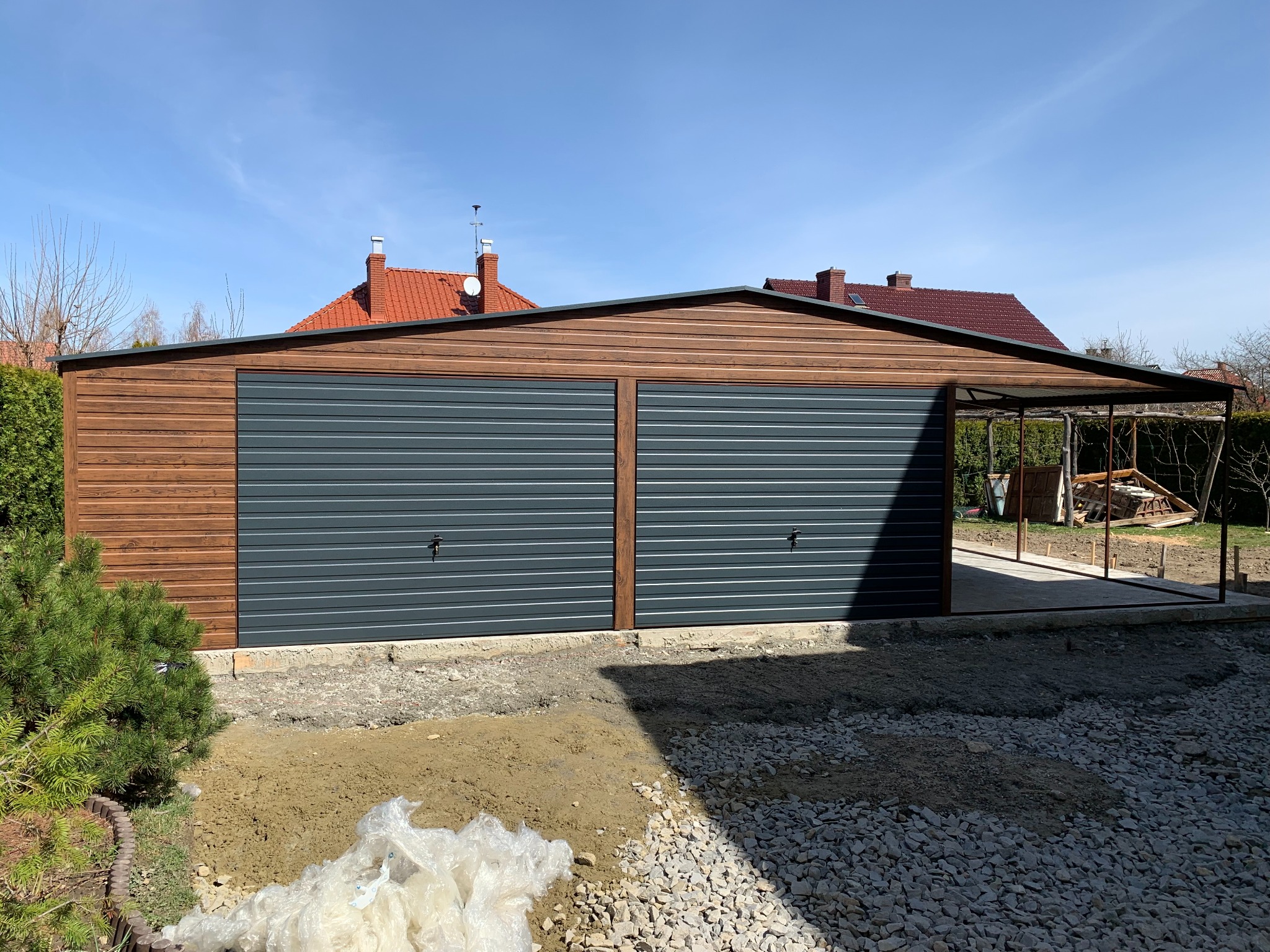 Nowoczesny garaż dwustanowiskowy z drewnianą elewacją i dwoma szarymi bramami segmentowymi, obok wiata. Widoczne przygotowane podłoże.