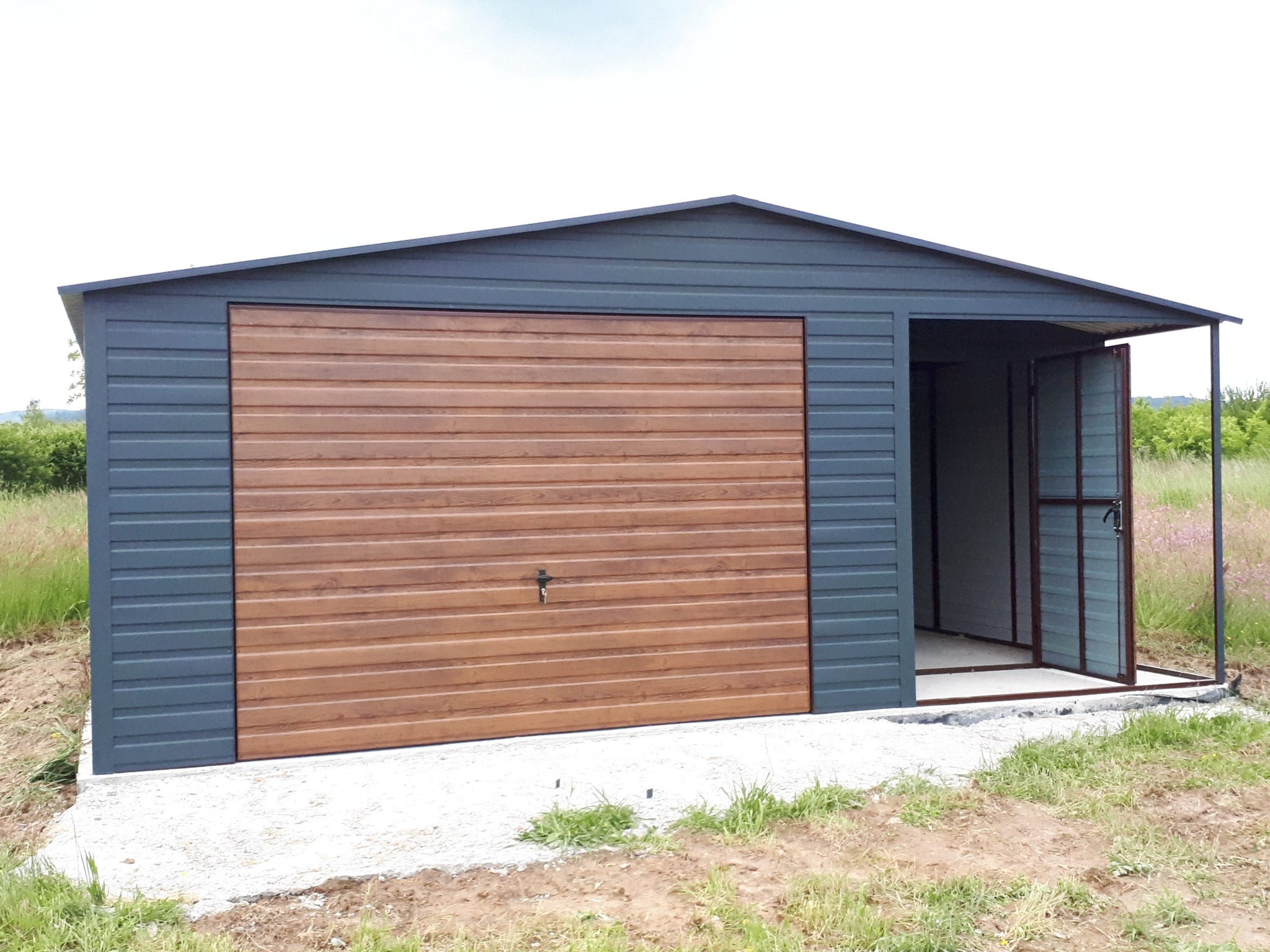 Szary garaż blaszany z bramą segmentową w kolorze drewna i otwartymi drzwiami bocznymi, postawiony na betonowej wylewce, na tle zieleni.