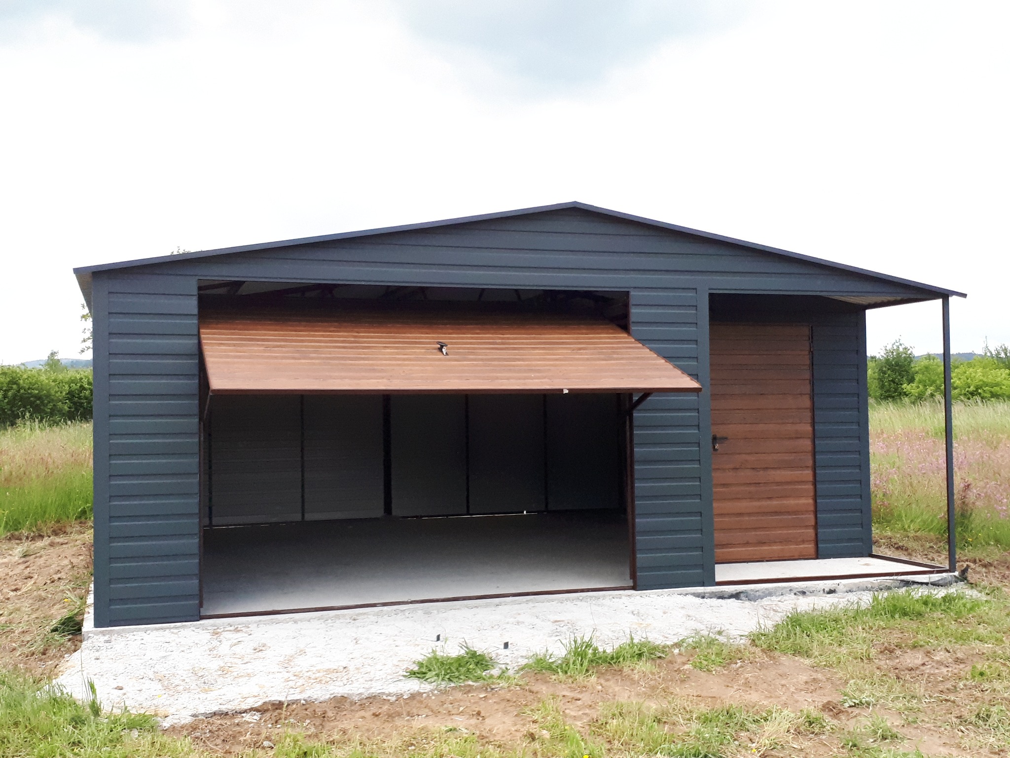 Szary garaż blaszany z otwartą bramą segmentową w kolorze drewna i drzwiami wejściowymi z boku, na betonowej wylewce, otoczony zielenią.