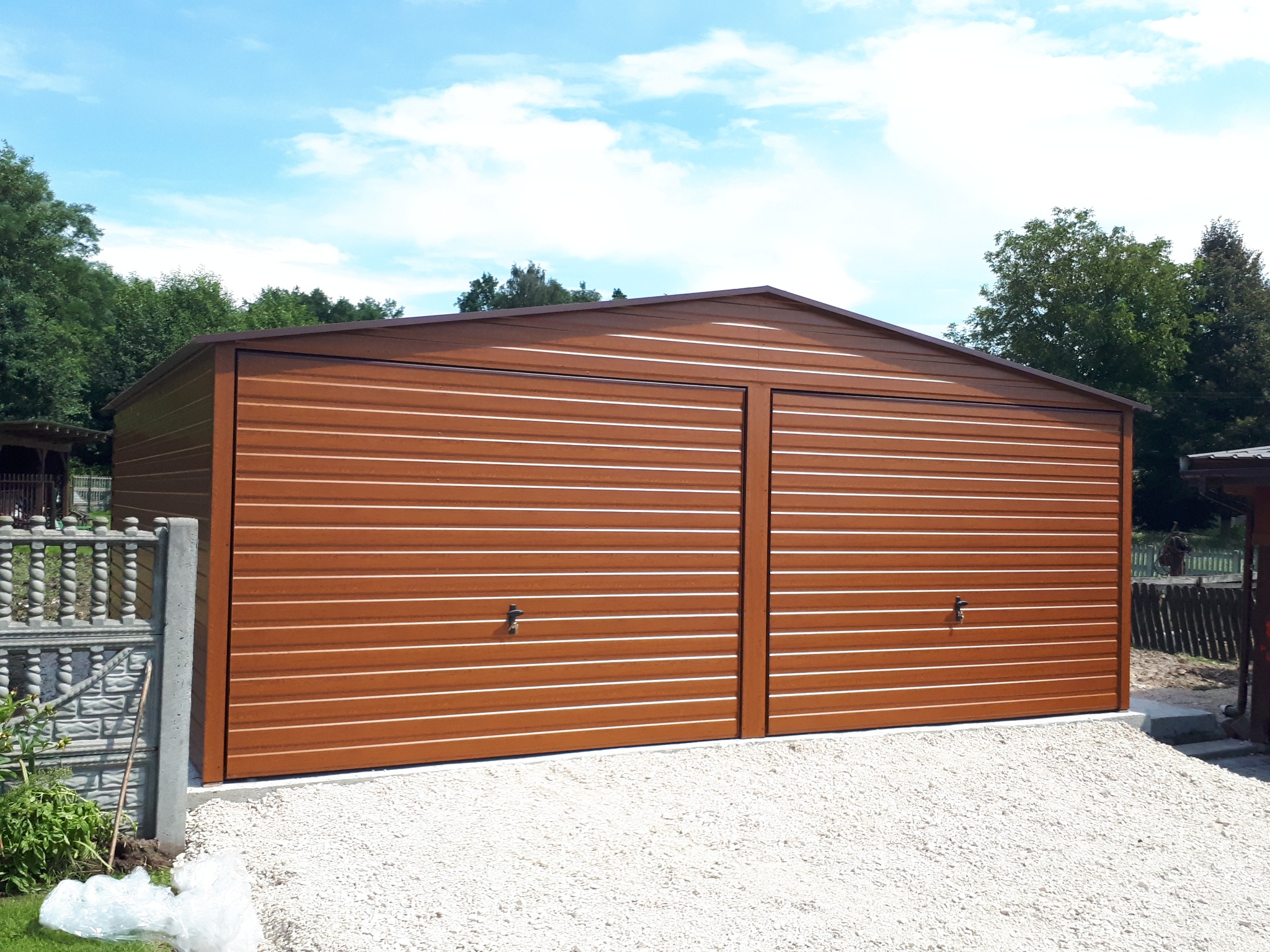 Dwustanowiskowy garaż blaszany w kolorze brązowym, z poziomymi przetłoczeniami, na podłożu z białego żwiru, w tle zieleń drzew i błękitne niebo.