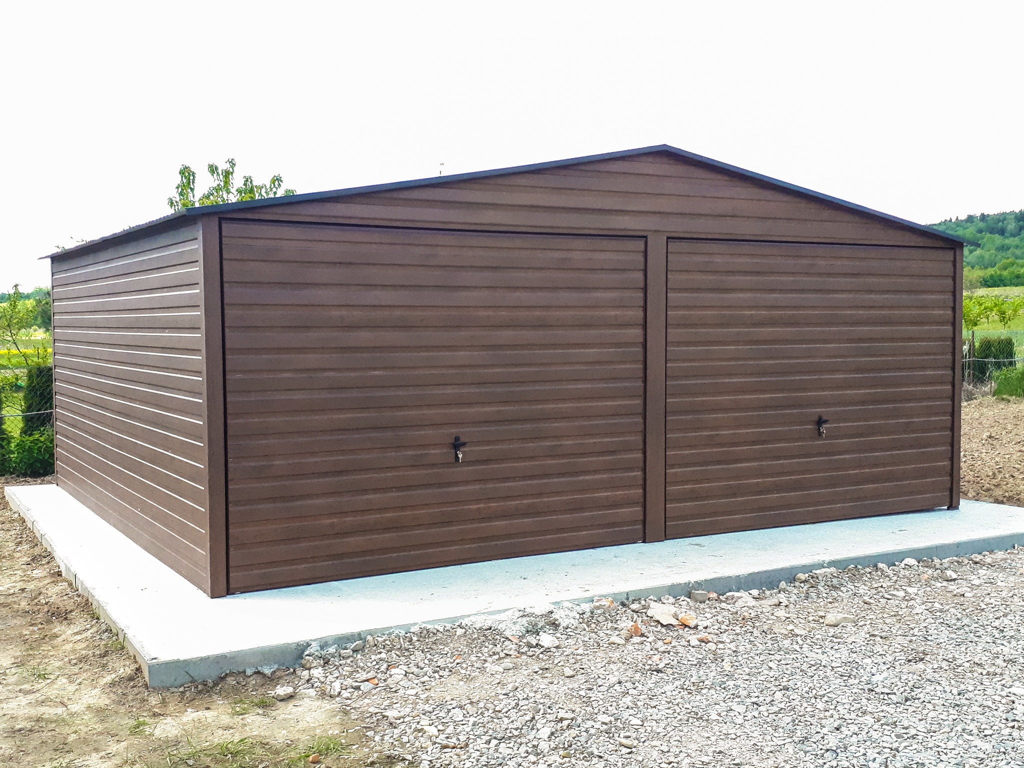 Brązowy garaż z dwoma bramami segmentowymi, zamykanymi na klucz, na betonowej wylewce, otoczony żwirem i zielenią.