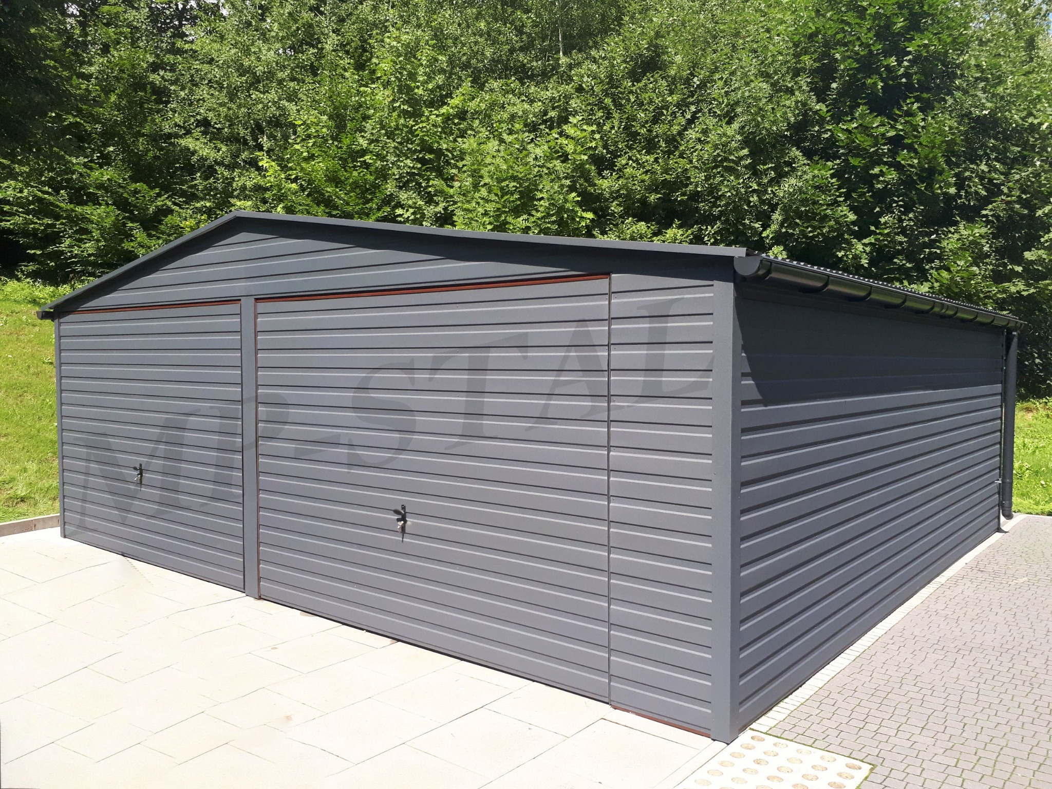 Szary garaż blaszany dwustanowiskowy z dachem dwuspadowym, widok z boku na tle zieleni, z widocznymi detalami jak rynna i faktura blachy.