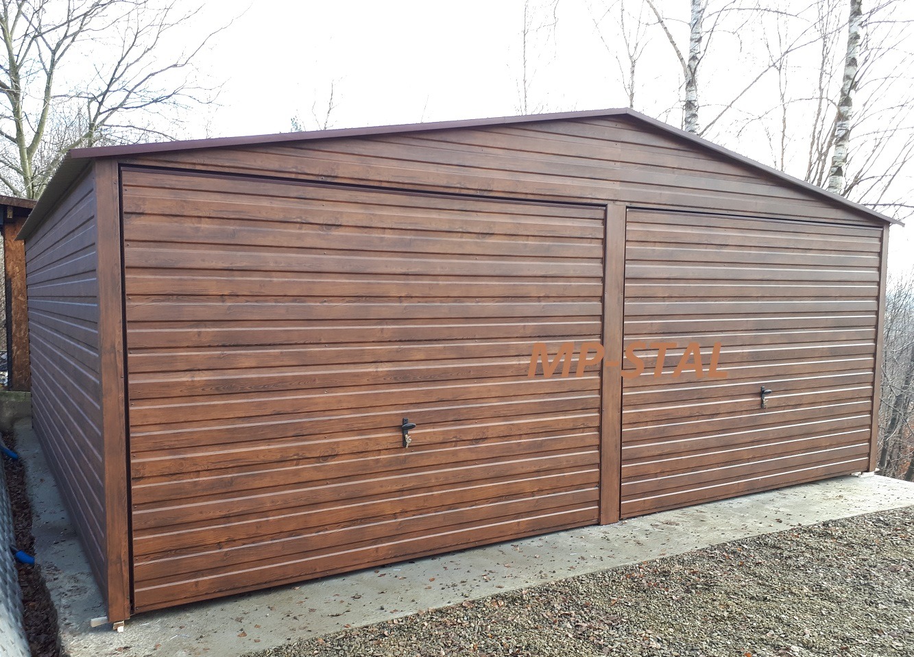 Dwustanowiskowy garaż blaszany w odcieniach brązu, z poziomymi panelami i dwuspadowym dachem, umieszczony na betonowej wylewce, na tle drzew bezlistnych.