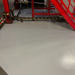 K - FLOOR Posadzki Żywicze Krzysztof Kudzia - Gładka, jasnoszara posadzka w hali przemysłowej, z widocznymi fragmentami czerwonej konstrukcji stalowej i taśm ostrzegawczych. Nowa, lśniąca powierzchnia.