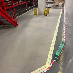 K - FLOOR Posadzki Żywicze Krzysztof Kudzia - Gładka, szara posadzka przemysłowa w magazynie, wyznaczona żółtą taśmą i czerwoną barierką. Widoczne elementy konstrukcyjne magazynu i palety w tle.