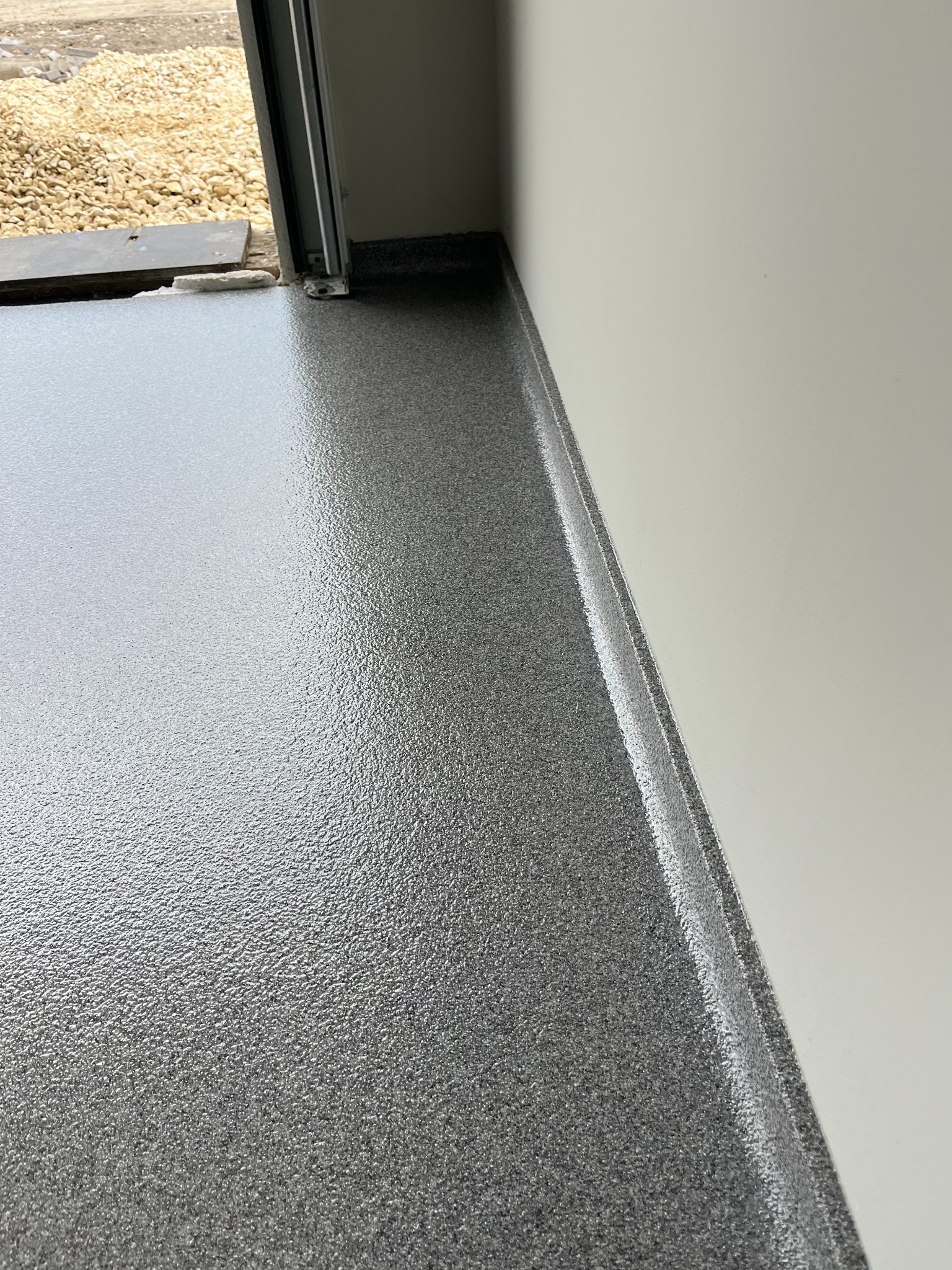 Szara, błyszcząca powierzchnia podłogi z widoczną fakturą, przylegająca do białej ściany i progu drzwi. Widoczne drobne kamienie na zewnątrz budynku.
