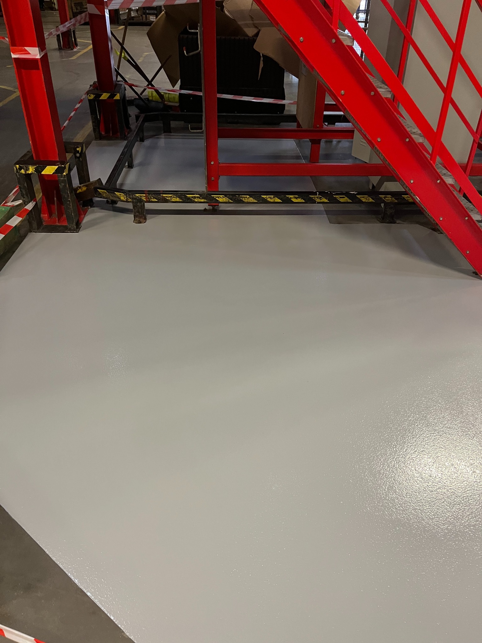 Gładka, jasnoszara posadzka w hali przemysłowej, z widocznymi fragmentami czerwonej konstrukcji stalowej i taśm ostrzegawczych. Nowa, lśniąca powierzchnia.