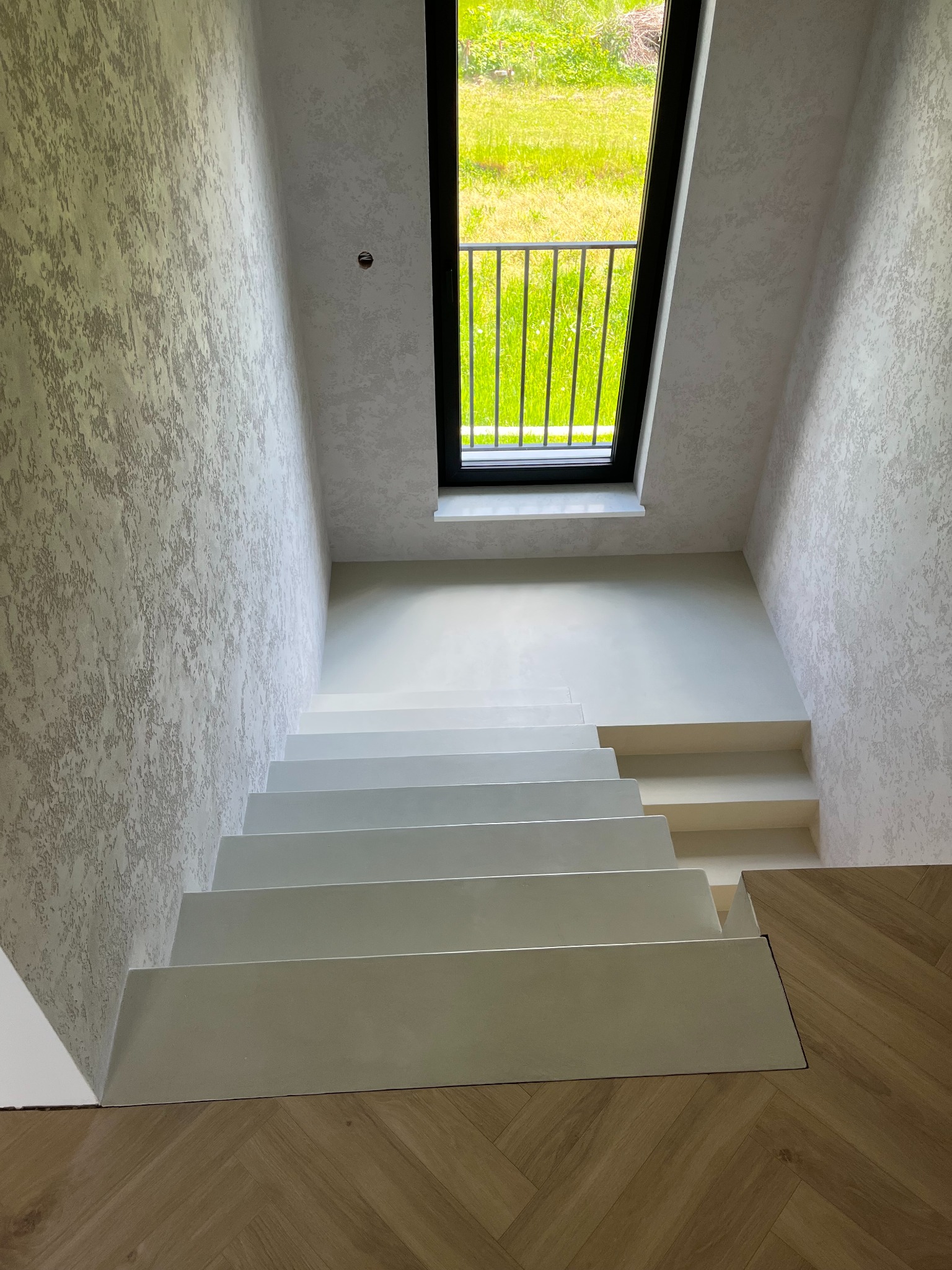 Nowoczesne schody z jasnego materiału prowadzą w dół, ściany pokryte dekoracyjnym tynkiem. Okno z widokiem na zieleń w tle. Minimalistyczny design.