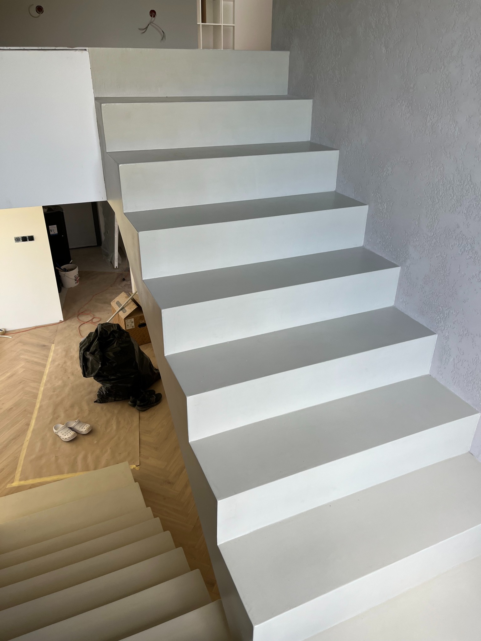 Białe schody z mikrocementu w nowoczesnym wnętrzu, widok z góry, ściana z efektem dekoracyjnym, zabezpieczona podłoga, etap prac wykończeniowych.