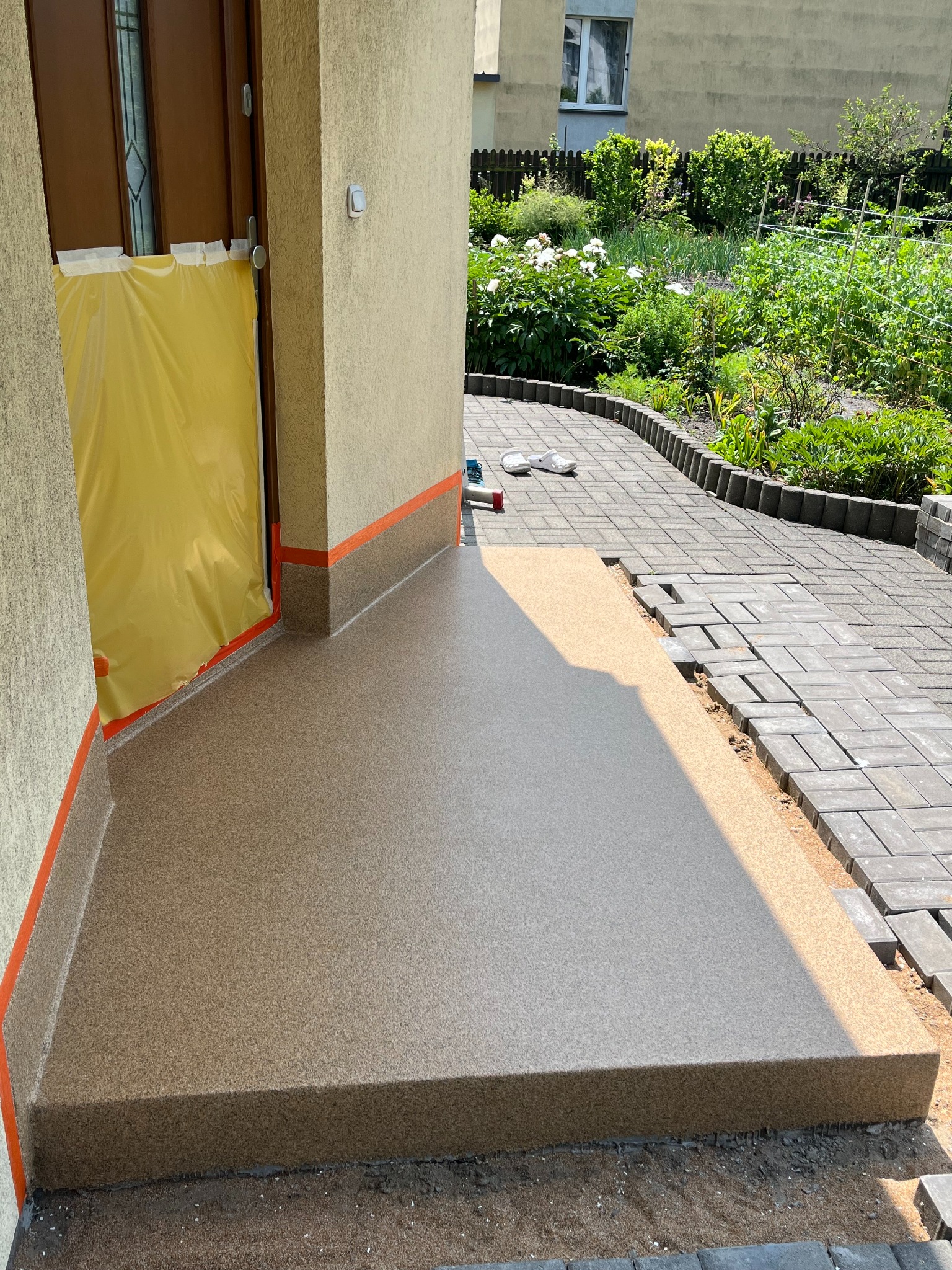 Wykończony taras z beżową, dekoracyjną powierzchnią. Wejście do domu zabezpieczone folią. Część tarasu w trakcie układania kostki brukowej na zewnątrz.