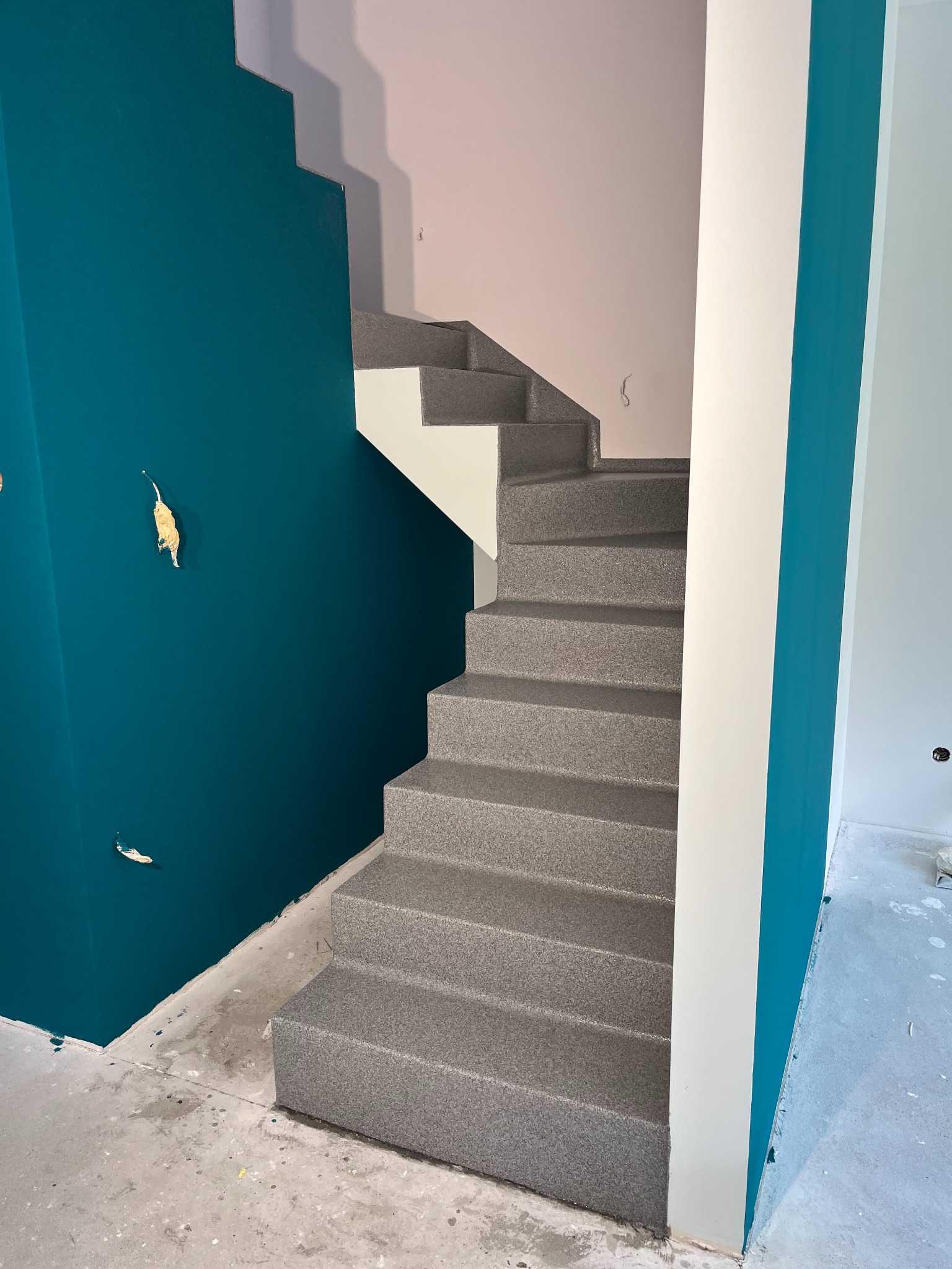 Schody pokryte szarą, dekoracyjną posadzką żywiczną. Ściana w odcieniach turkusu i bieli. Widoczne ślady po taśmie malarskiej na ścianie.