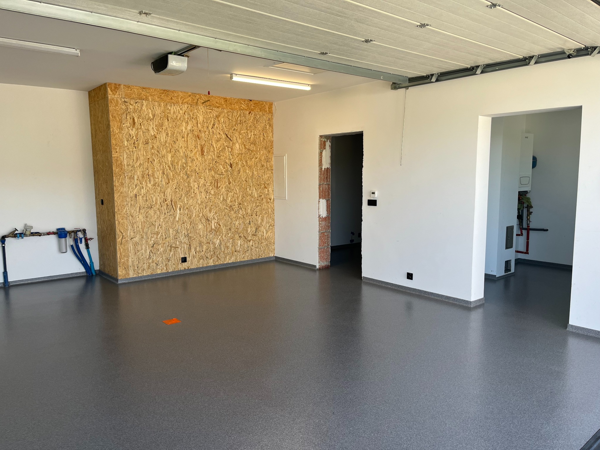 Szara, gładka posadzka w garażu z widocznymi instalacjami przy ścianie. Ściany białe, fragment ściany z płyt OSB. Widoczne drzwi do pomieszczenia.