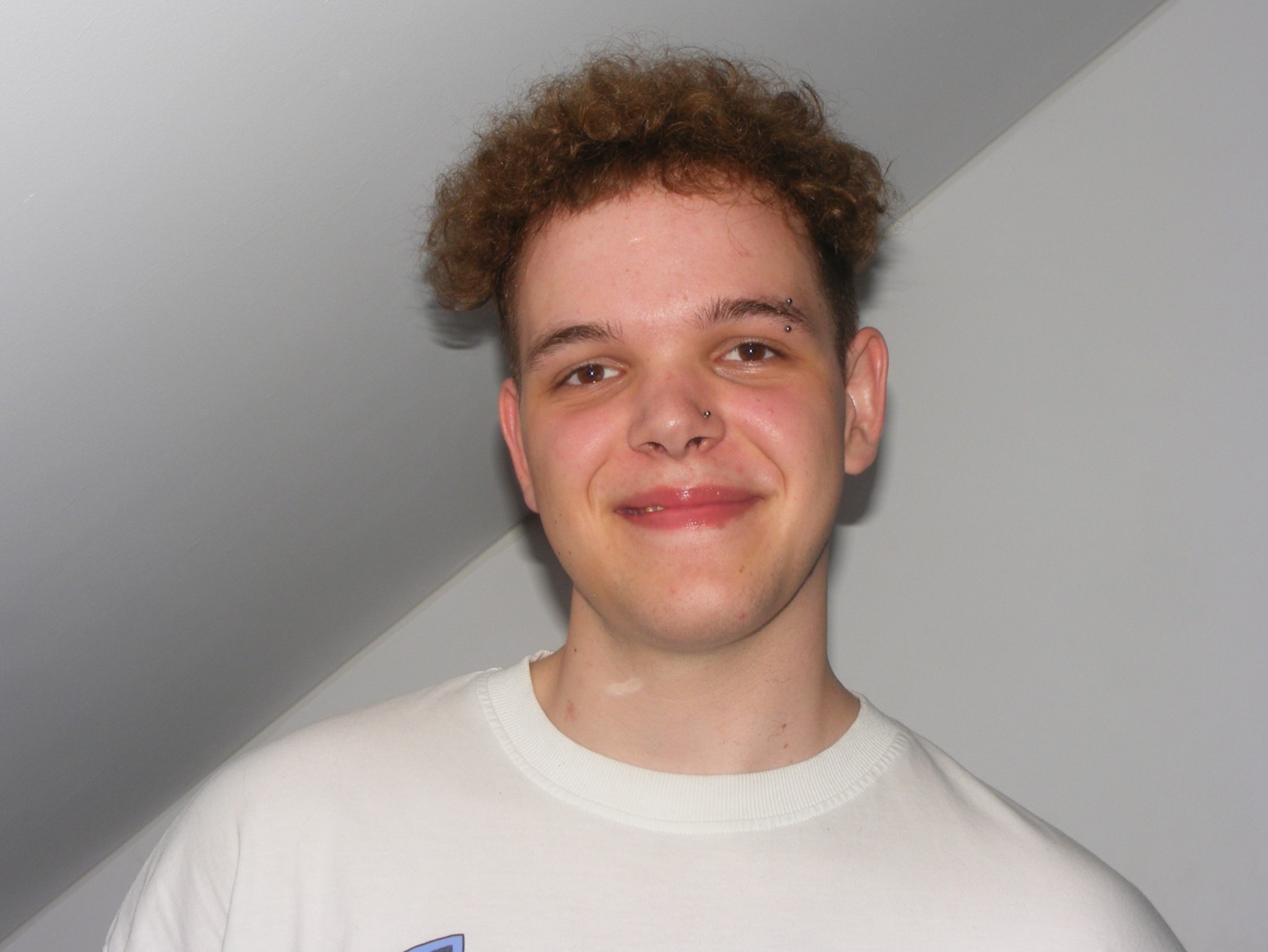 Portret uśmiechniętego młodego mężczyzny z kręconymi włosami i piercingiem brwi oraz nosa, na tle jasnej ściany. Widoczna biała koszulka.