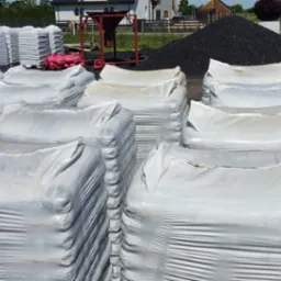 Stosy pelletu drzewnego w foliowych workach na paletach, z kopcem węgla w tle, na placu składowym. Widoczny wózek widłowy i sprzęt do załadunku.