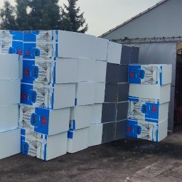 PHU Bartosz Gruszka - Ułożone kostki styropianu EPS z niebieskim nadrukiem, tworzące piramidę na zewnątrz magazynu budowlanego. Widoczne narzędzia i zieleń w tle.