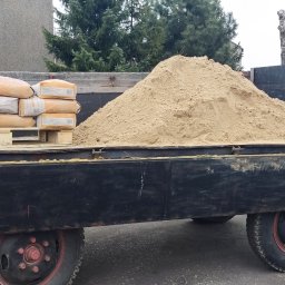 PHU Bartosz Gruszka - Ciężarówka z ładunkiem: piasek obok palety z workami cementu, na tle hali magazynowej i zieleni. Widoczne detale ciężarówki i faktura materiałów.