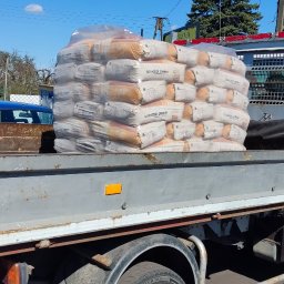 PHU Bartosz Gruszka - Ciężarówka z workami cementu marki Górażdże Cement Master na skrzyni ładunkowej. Widoczne numery kontaktowe na kabinie i otoczenie składu budowlanego.