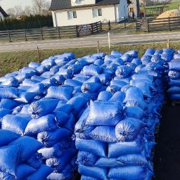 PHU Bartosz Gruszka - Stosy niebieskich worków na paletach, z czerwoną konstrukcją zsypu w tle, na tle zabudowań i drogi. Widoczny znak reklamowy w oddali.