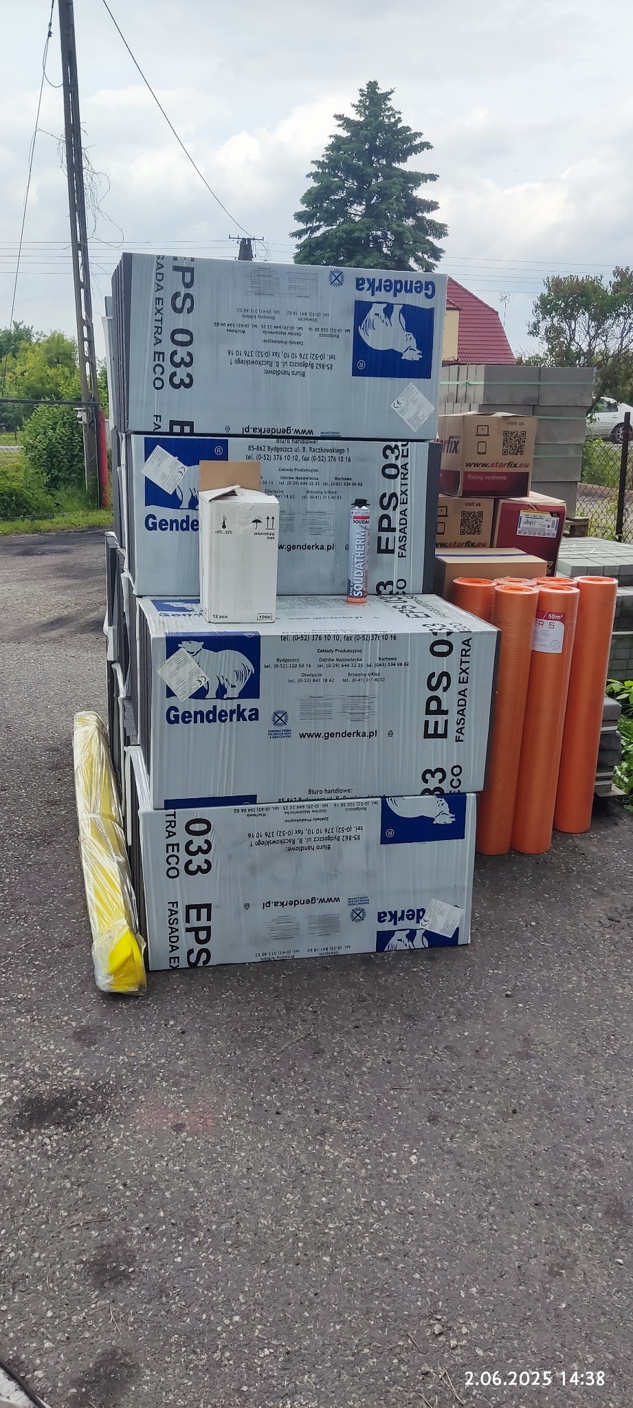 Styropian fasadowy Genderka EPS 033 ułożony w stos, obok rolki folii i piana montażowa. Widoczne opakowania zaprawy murarskiej i kostka brukowa w tle.