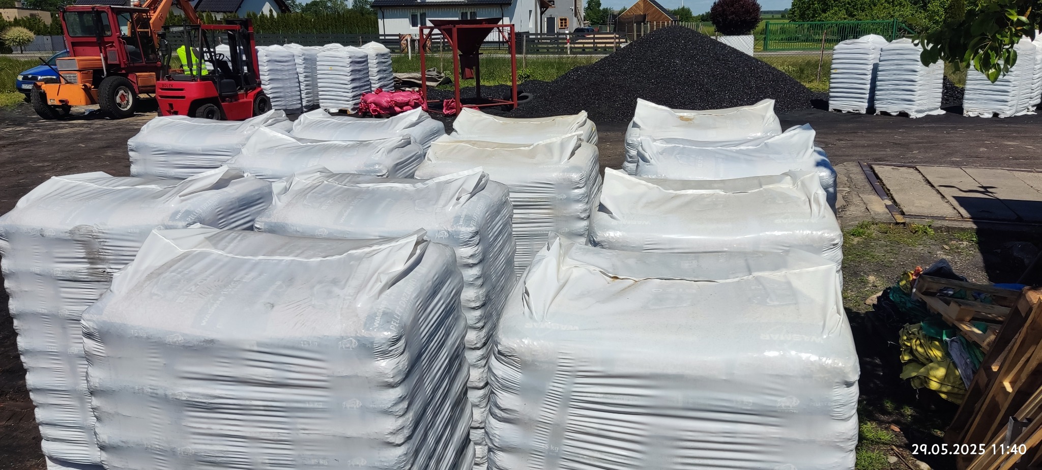 Stosy pelletu drzewnego w foliowych workach na paletach, z kopcem węgla w tle, na placu składowym. Widoczny wózek widłowy i sprzęt do załadunku.