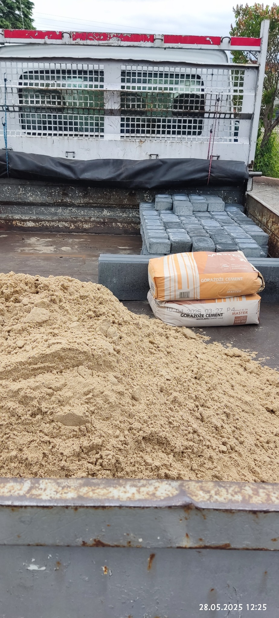 Ładunek na pace samochodu: piasek, kostka brukowa i dwa worki cementu Gorazdze Master, gotowe do transportu na budowę lub do klienta.