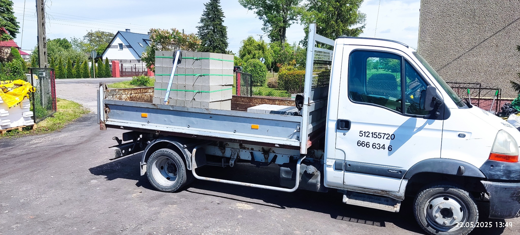 Ciężarówka z platformą załadowana betonowymi bloczkami, zabezpieczonymi taśmami. Widoczne numery kontaktowe na kabinie i otoczenie: droga, budynki, zieleń.