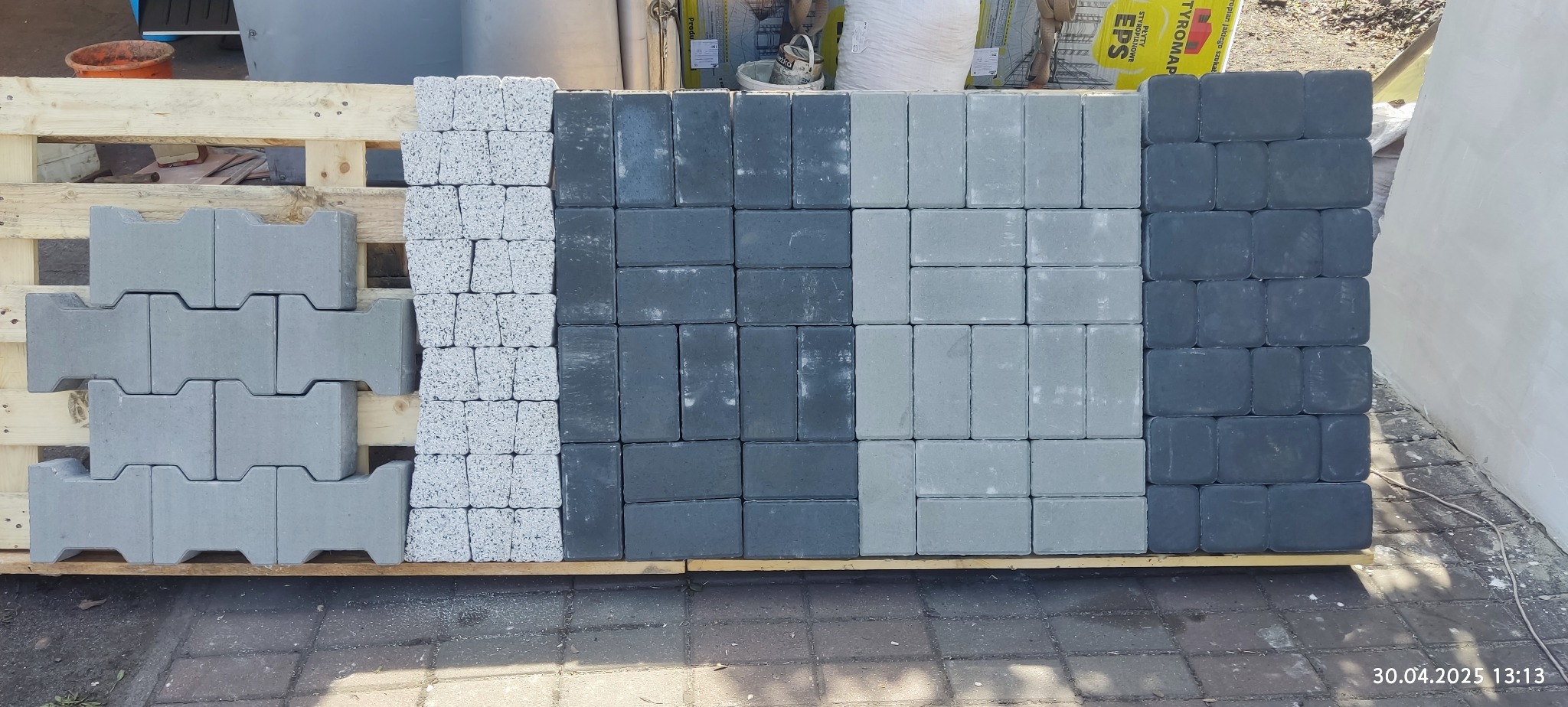 Paleta z kostką brukową w różnych odcieniach szarości i kształtach, ułożona rzędami. Widoczne faktury i detale materiału budowlanego.