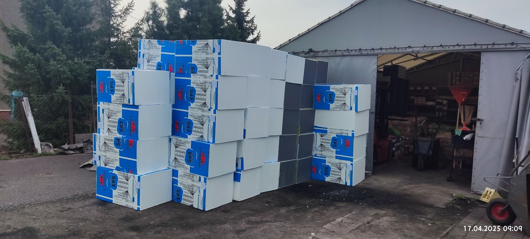 Ułożone kostki styropianu EPS z niebieskim nadrukiem, tworzące piramidę na zewnątrz magazynu budowlanego. Widoczne narzędzia i zieleń w tle.