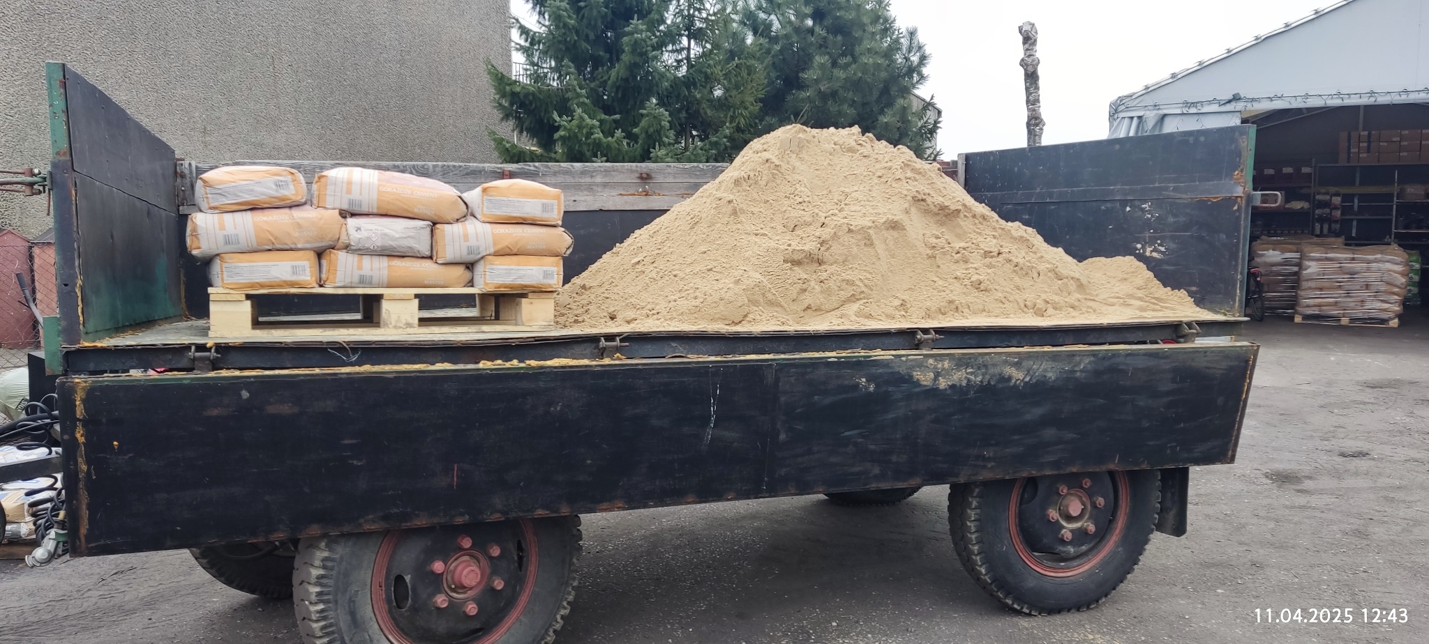 Ciężarówka z ładunkiem: piasek obok palety z workami cementu, na tle hali magazynowej i zieleni. Widoczne detale ciężarówki i faktura materiałów.