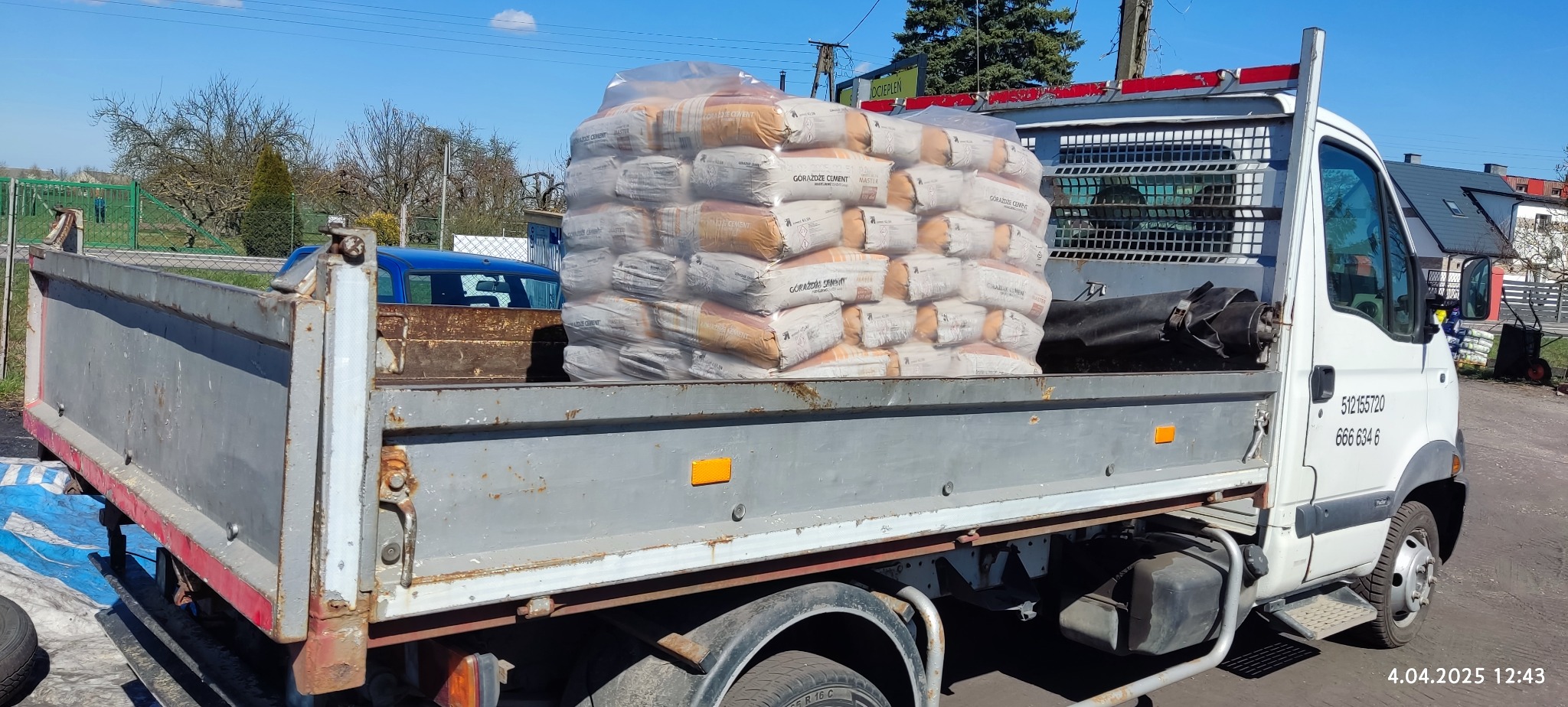 Ciężarówka z workami cementu marki Górażdże Cement Master na skrzyni ładunkowej. Widoczne numery kontaktowe na kabinie i otoczenie składu budowlanego.