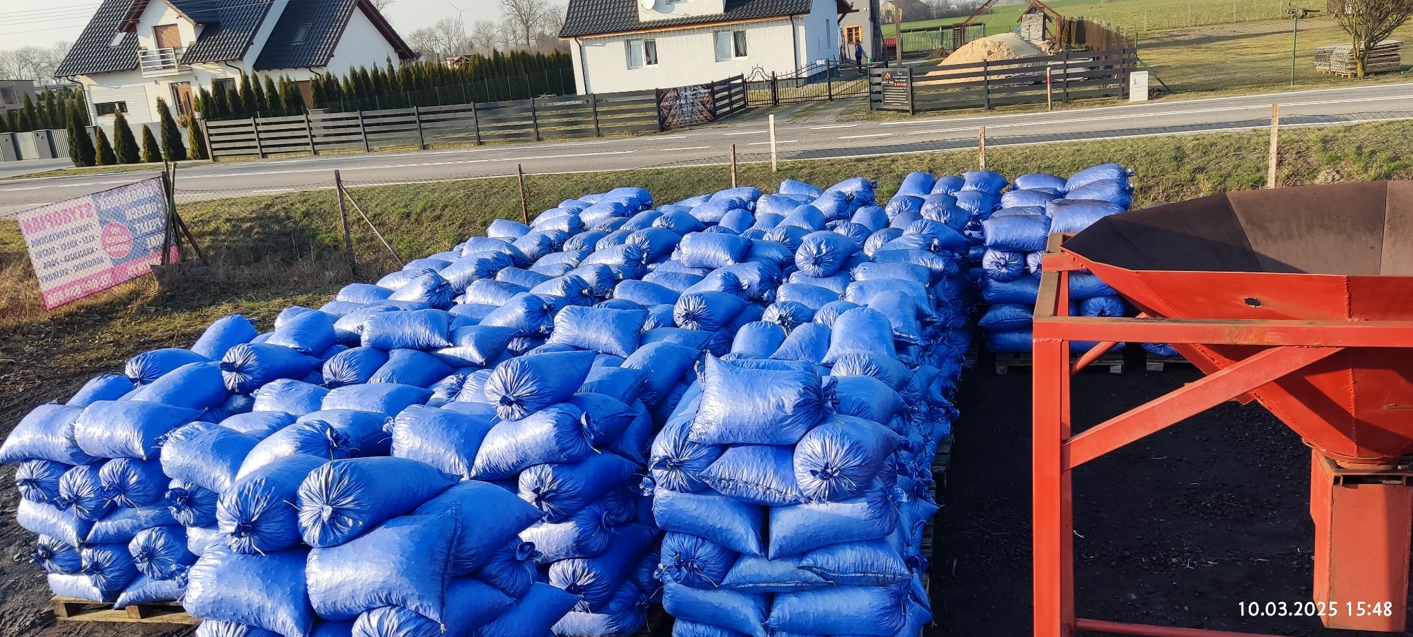 Stosy niebieskich worków na paletach, z czerwoną konstrukcją zsypu w tle, na tle zabudowań i drogi. Widoczny znak reklamowy w oddali.