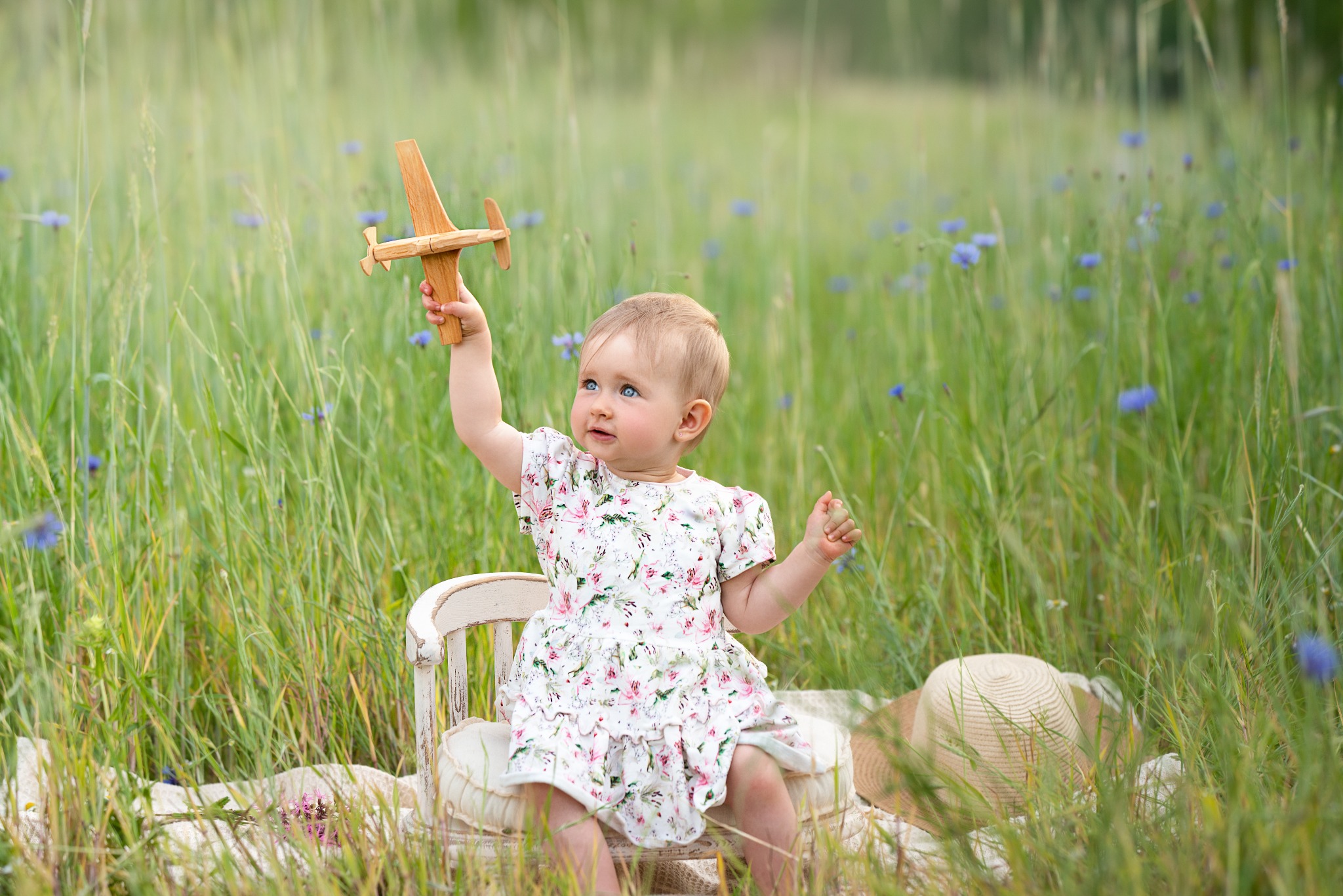 Urocza blondwłosa dziewczynka w kwiecistej sukience siedzi na krześle w wysokiej trawie, trzymając drewniany model samolotu. W tle błękitne kwiaty polne.