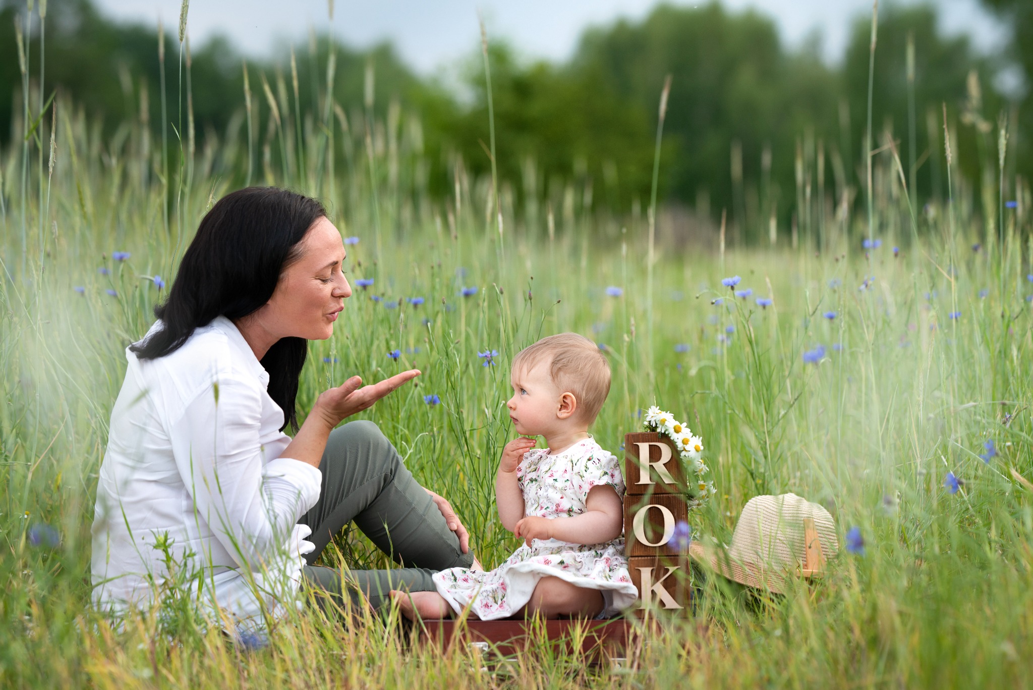 Sesja w plenerze: Mama w białej koszuli i córka w kwiecistej sukience siedzą na łące pełnej błękitnych kwiatów, obok drewniane klocki z napisem ROK i kapelusz.