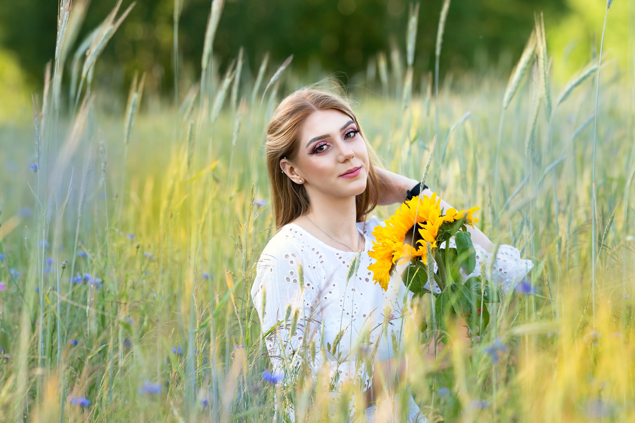 Portret kobiety w białej bluzce, trzymającej bukiet słoneczników na łące. Delikatny makijaż, naturalne światło, subtelny uśmiech. Romantyczna sesja w plenerze.
