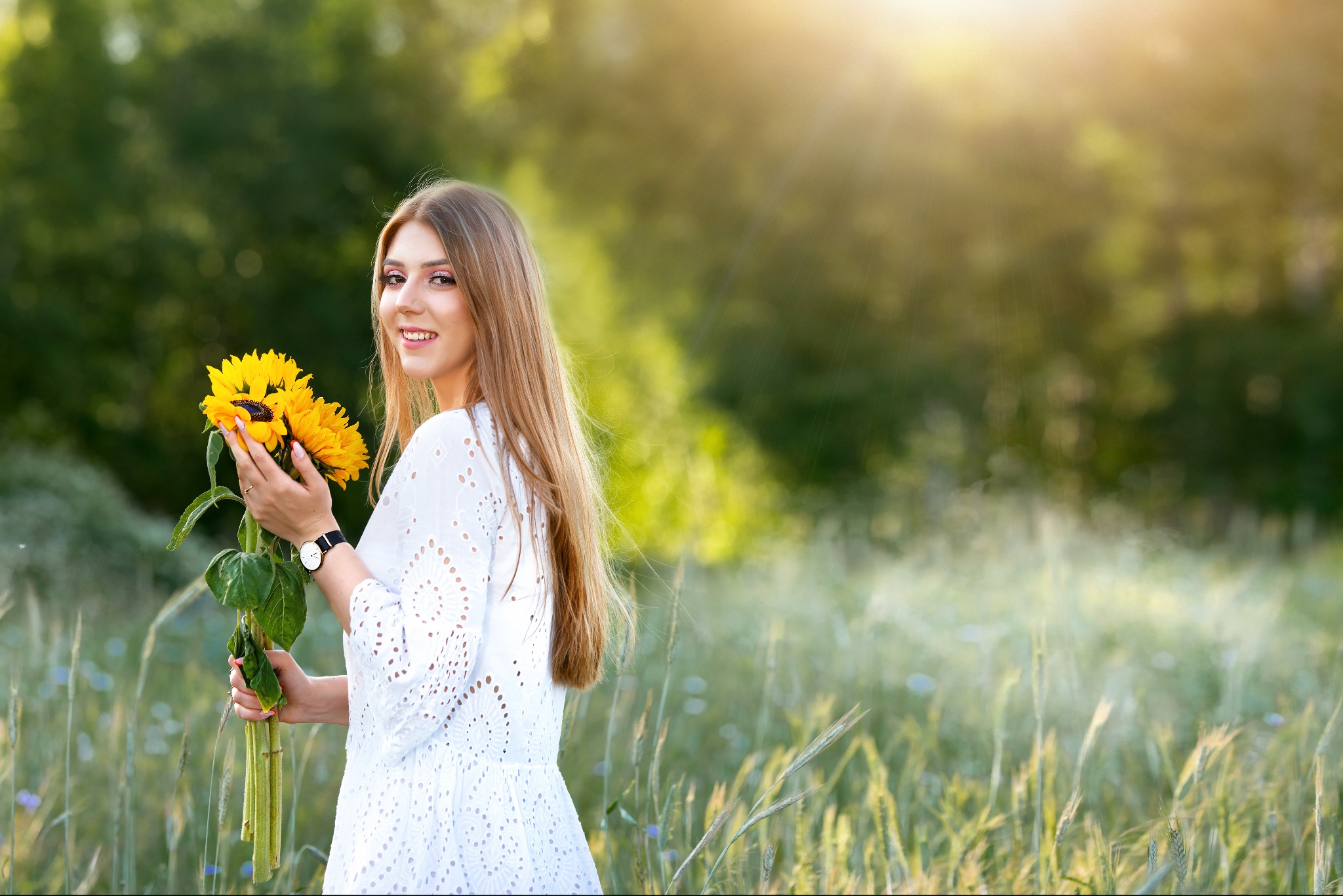 Uśmiechnięta kobieta w białej sukience, trzymająca bukiet słoneczników na łące w promieniach słońca. Widoczny zegarek na ręce i delikatny makijaż.
