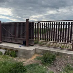 Przesuwna brama z automatyką w kolorze brązowym na betonowej podstawie, widok z zewnątrz, pochmurne niebo w tle i usypany kopiec kamieni.