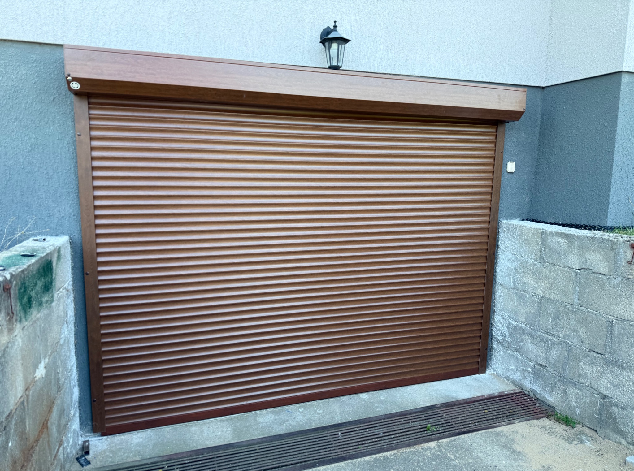 Zewnętrzna brama rolowana w kolorze brązowym, zamontowana w garażu. Nad bramą widoczna lampa zewnętrzna. Szara ściana budynku i betonowe obramowanie bramy.