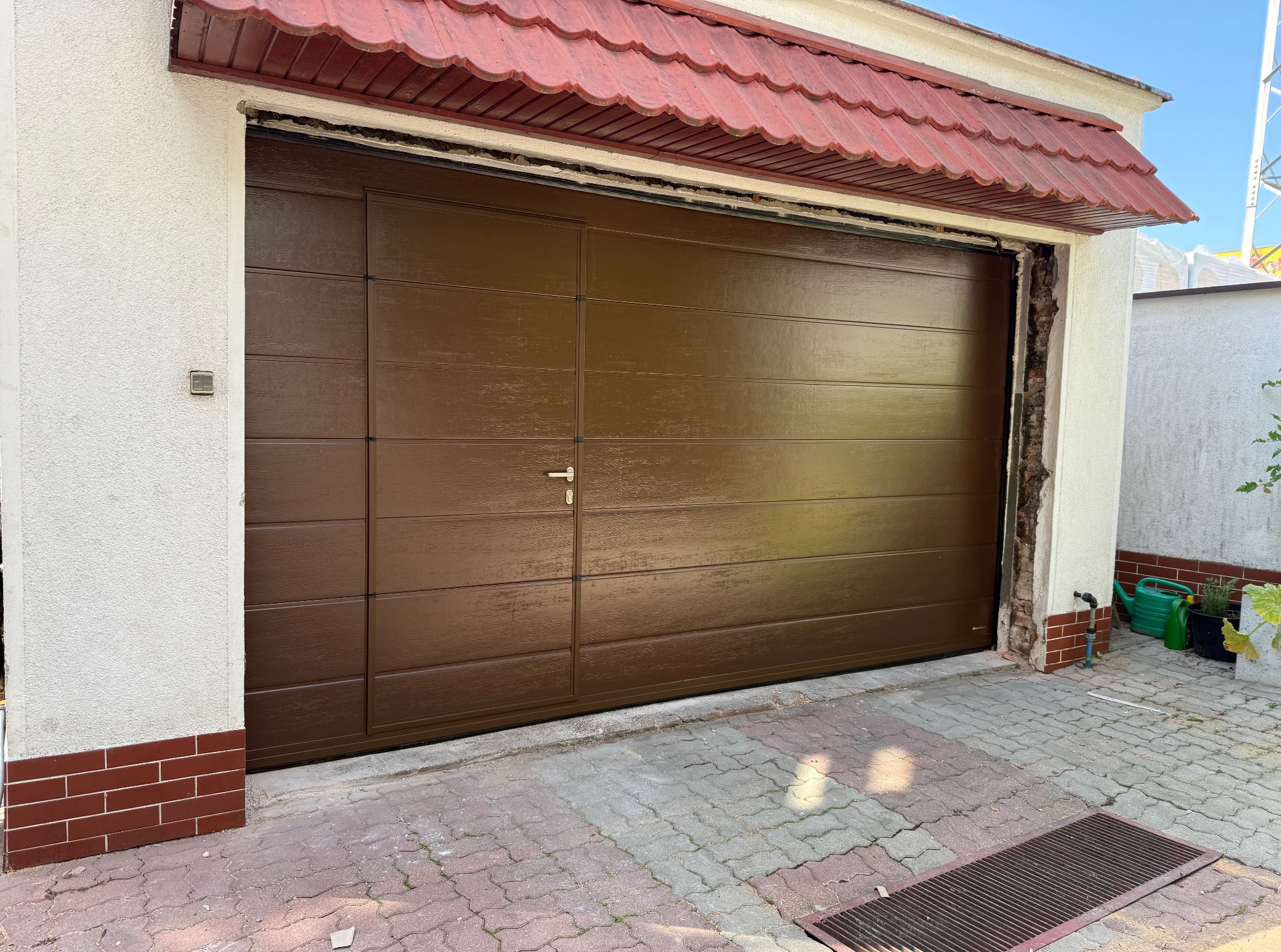 Brązowa segmentowa brama garażowa z poziomymi panelami, zamontowana w białym budynku z czerwonym dachem. Widoczne detale wykończenia i kostka brukowa przed garażem.