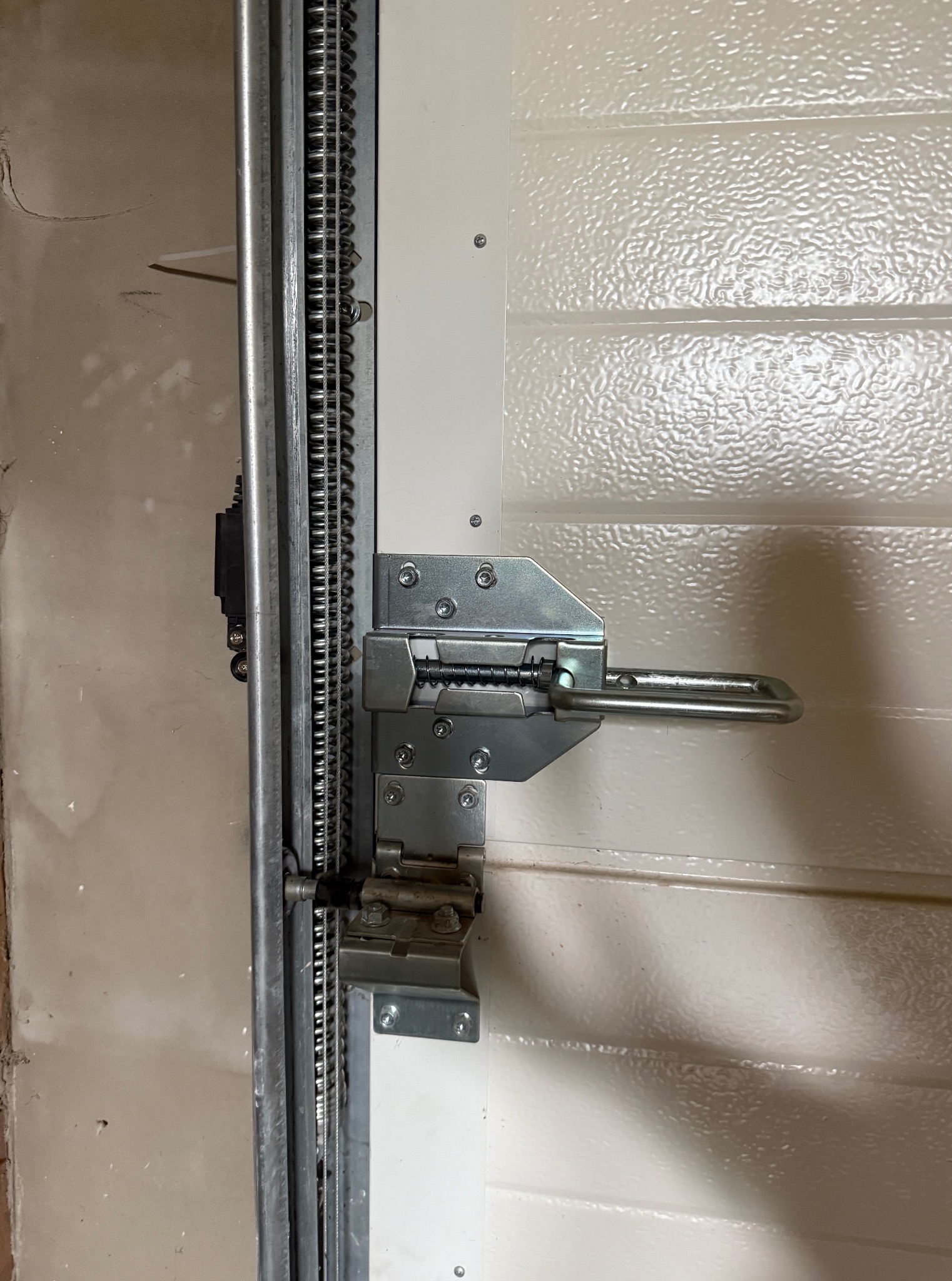 Szczegół mechanizmu bramy garażowej: łańcuch, prowadnica i metalowe mocowania na teksturowanym, białym panelu bramy. Widoczne ślady montażu na ścianie.