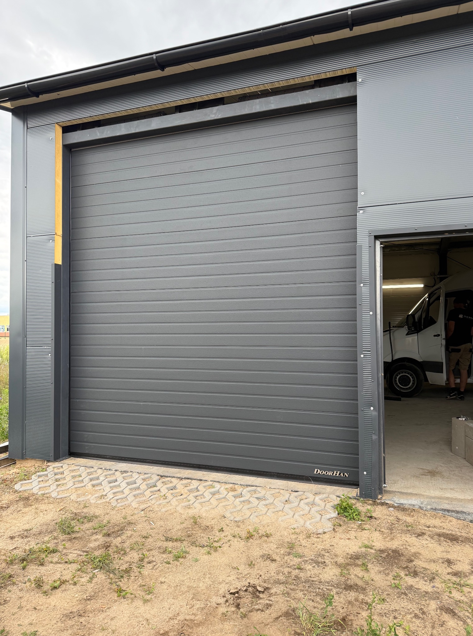 Szara brama segmentowa DoorHan w hali przemysłowej, widok z zewnątrz. W tle widać wnętrze hali z zaparkowanym białym busem.