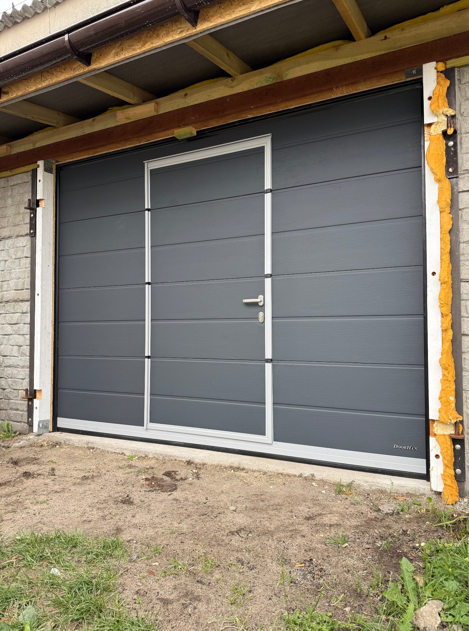 Szara segmentowa brama garażowa z drzwiami, widok z zewnątrz. Białe obramowanie, pianka montażowa przy murze. Brama marki DoorHan.