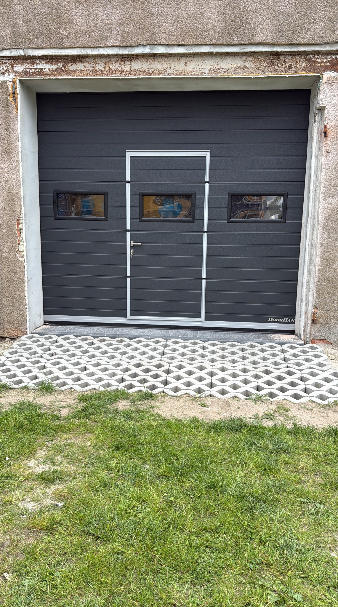 Szara segmentowa brama garażowa DoorHan z trzema małymi oknami i drzwiami wejściowymi, przed nią ażurowe płyty betonowe na trawniku.