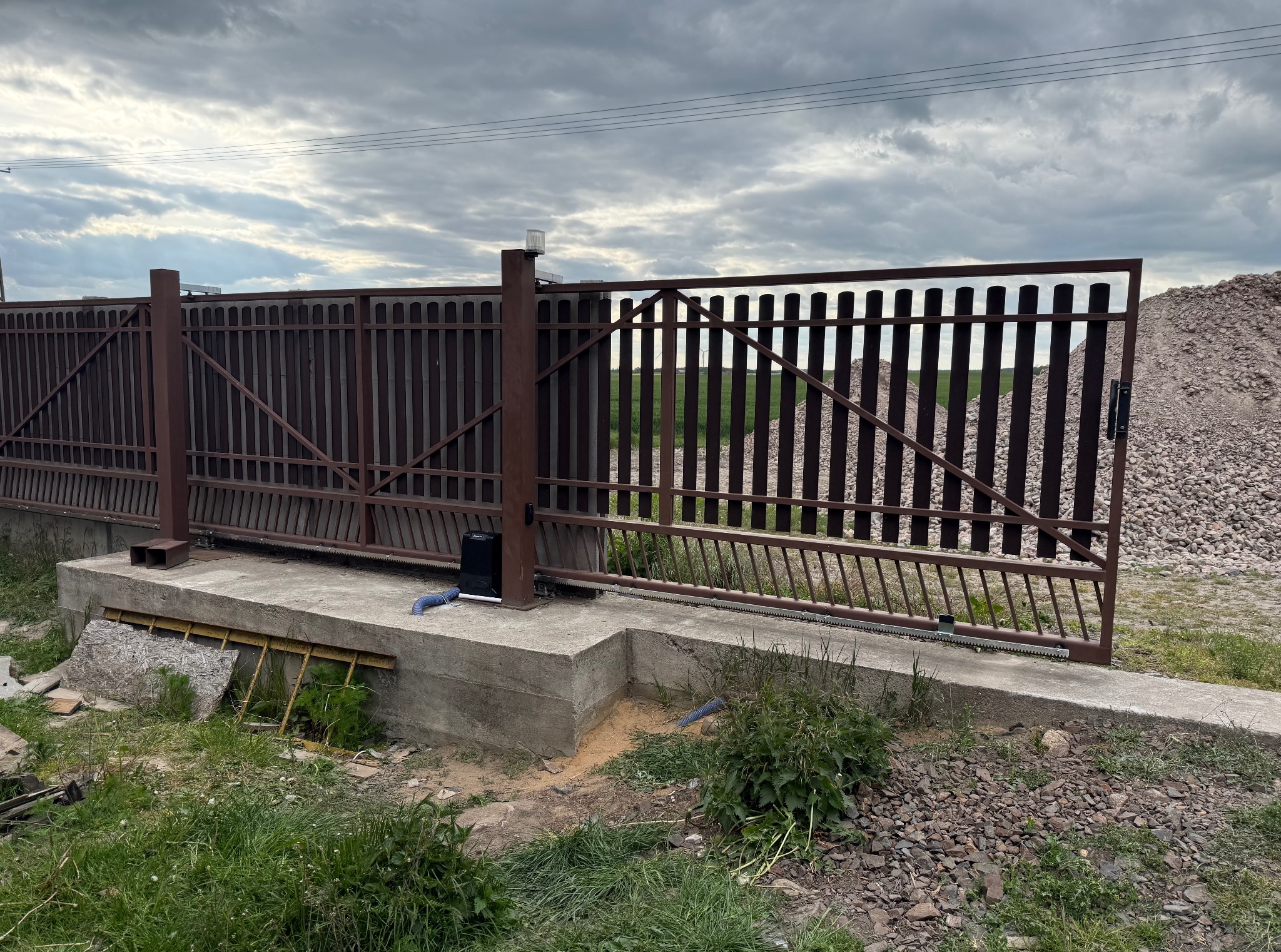 Przesuwna brama z automatyką w kolorze brązowym na betonowej podstawie, widok z zewnątrz, pochmurne niebo w tle i usypany kopiec kamieni.