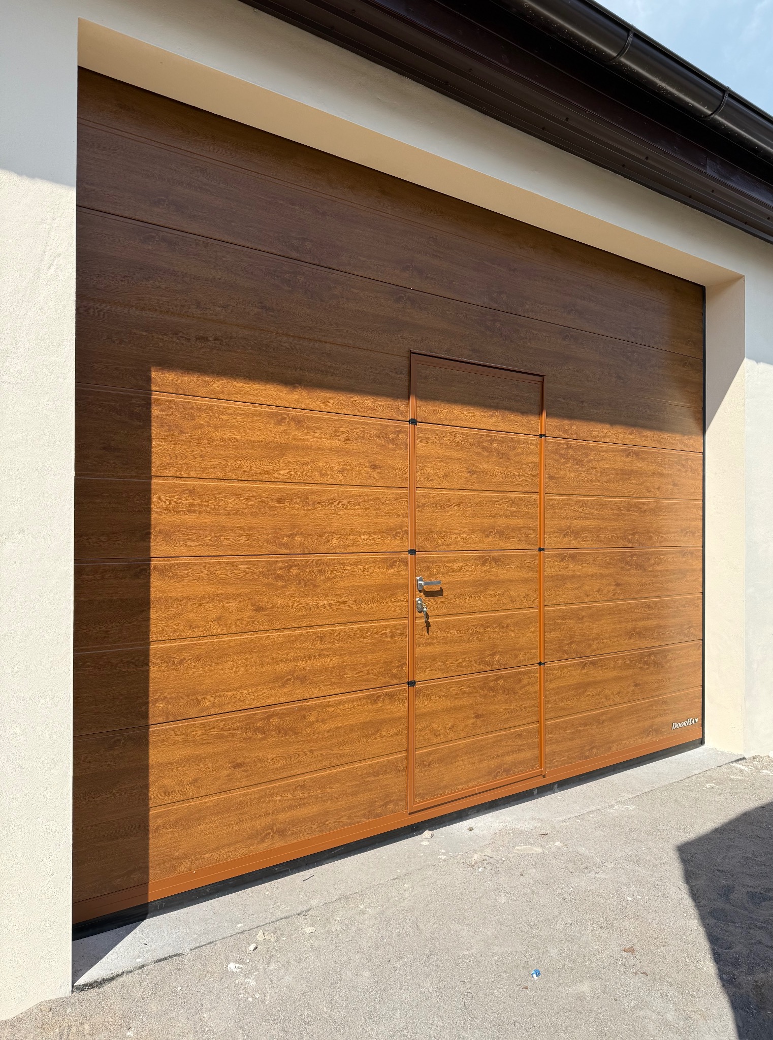 Brązowa brama garażowa segmentowa z drzwiami wejściowymi, klucz w zamku, imitacja drewna, widoczne detale paneli i uszczelnień.