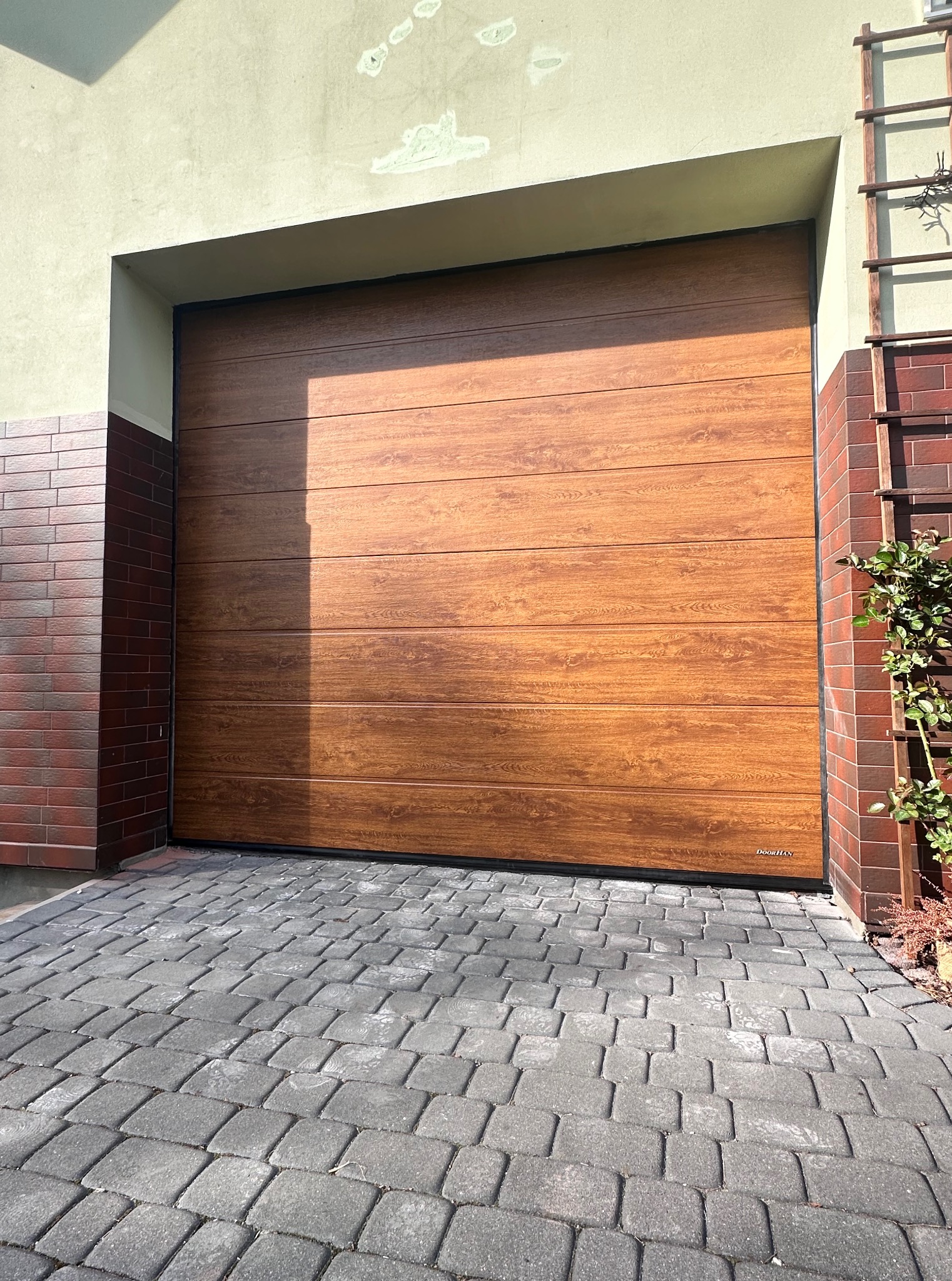 Zamknięta segmentowa brama garażowa w kolorze drewna, z widoczną fakturą paneli. Brama marki DoorHan, zamontowana w budynku z ceglaną elewacją i kostką brukową.