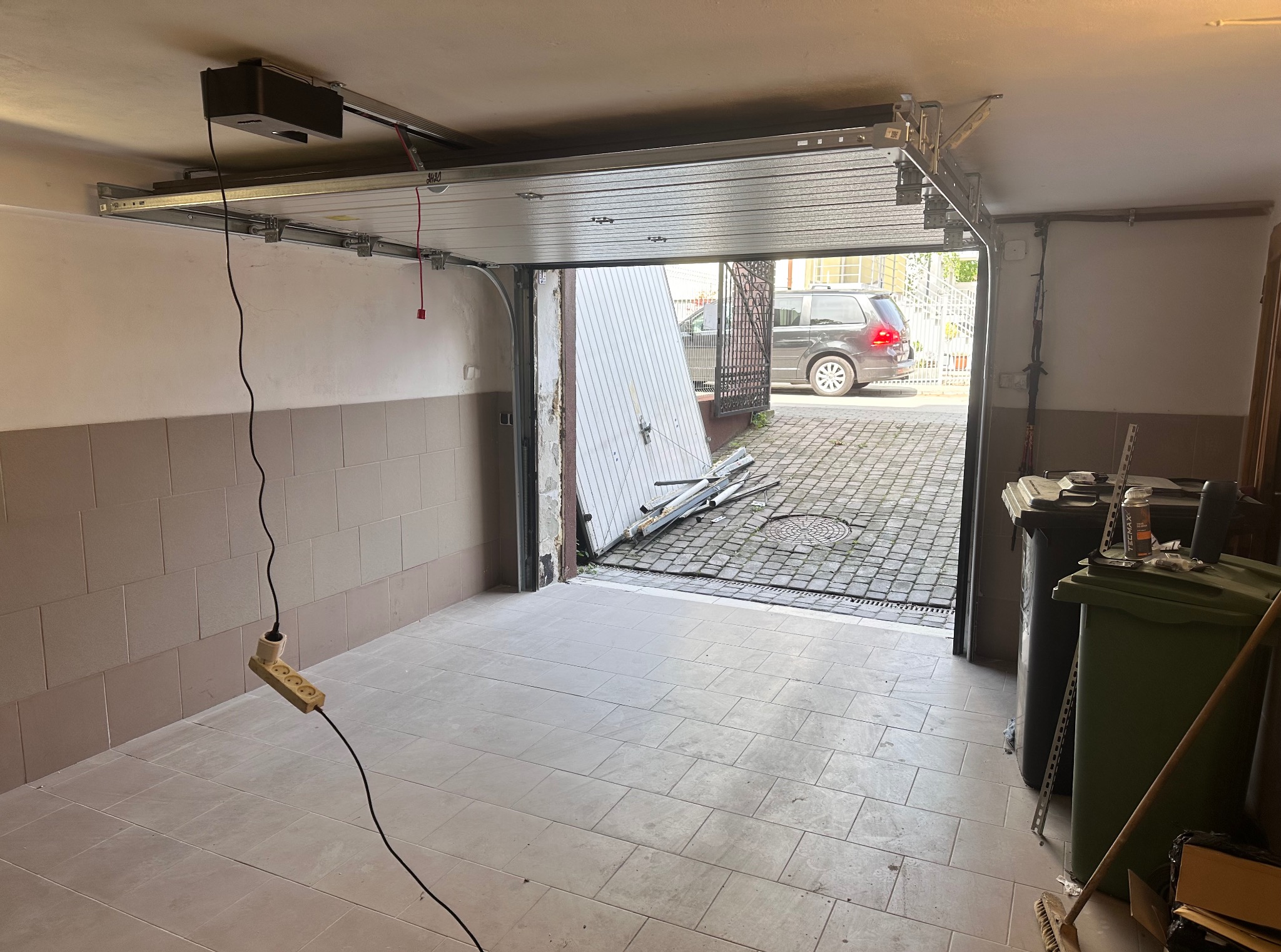 Wnętrze garażu z częściowo otwartą, segmentową bramą garażową w trakcie montażu. Widoczny napęd bramy, prowadnice i fragmenty paneli bramy. Na zewnątrz zaparkowany samochód.