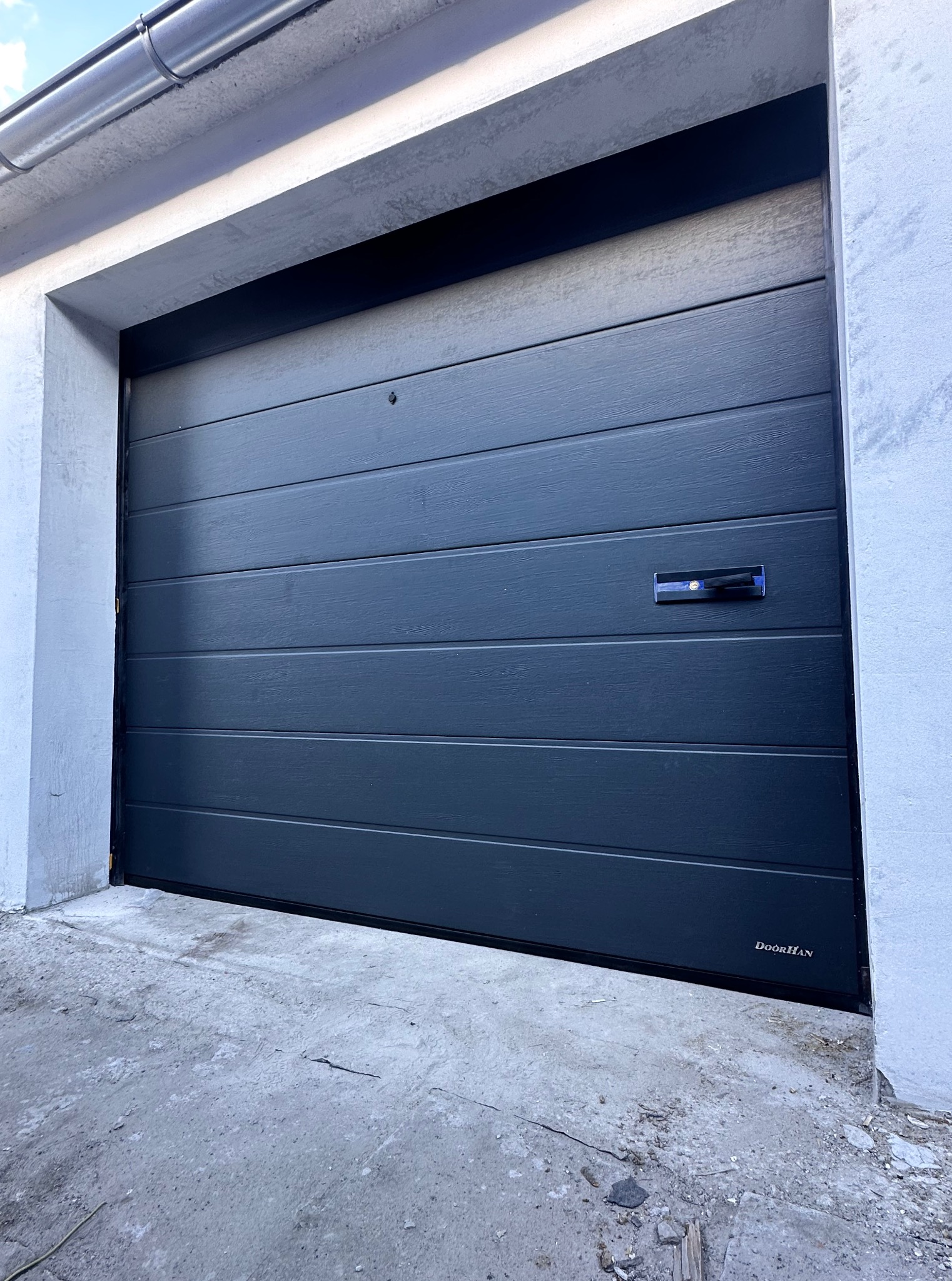 Grafitowa brama garażowa segmentowa DoorHan z poziomymi przetłoczeniami, zamontowana w surowym otworze garażowym. Widoczne zarysowania na posadzce.