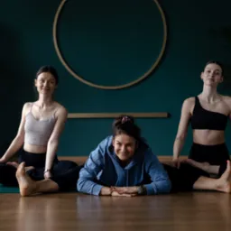 Trzy kobiety w strojach sportowych wykonują ćwiczenia jogi na matach, na tle ściany w kolorze butelkowej zieleni z dekoracyjnym złotym kołem.