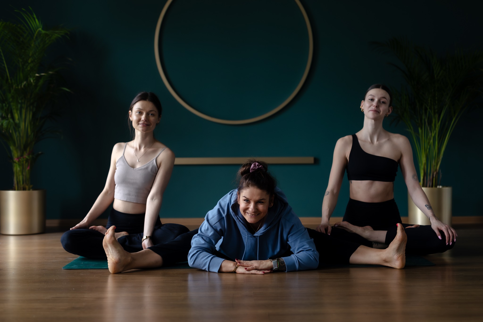 Trzy kobiety w strojach sportowych wykonują ćwiczenia jogi na matach, na tle ściany w kolorze butelkowej zieleni z dekoracyjnym złotym kołem.