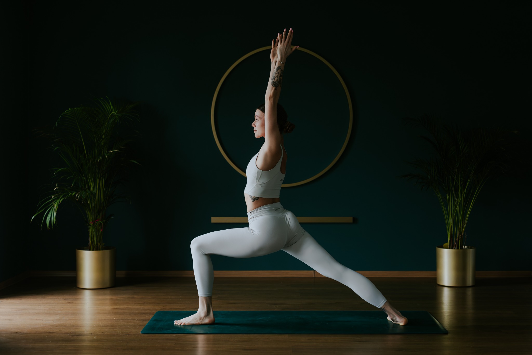 Kobieta w białym stroju wykonuje asanę wojownika I na macie do jogi, na tle ciemnozielonej ściany z okrągłą dekoracją.